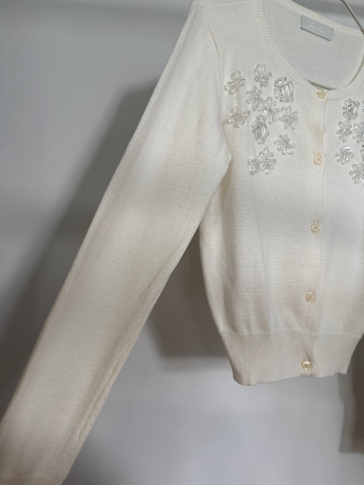 An off-white knit cardigan sweater with floral embellishments and buttons on the chest, shown hanging against a neutral gray background.