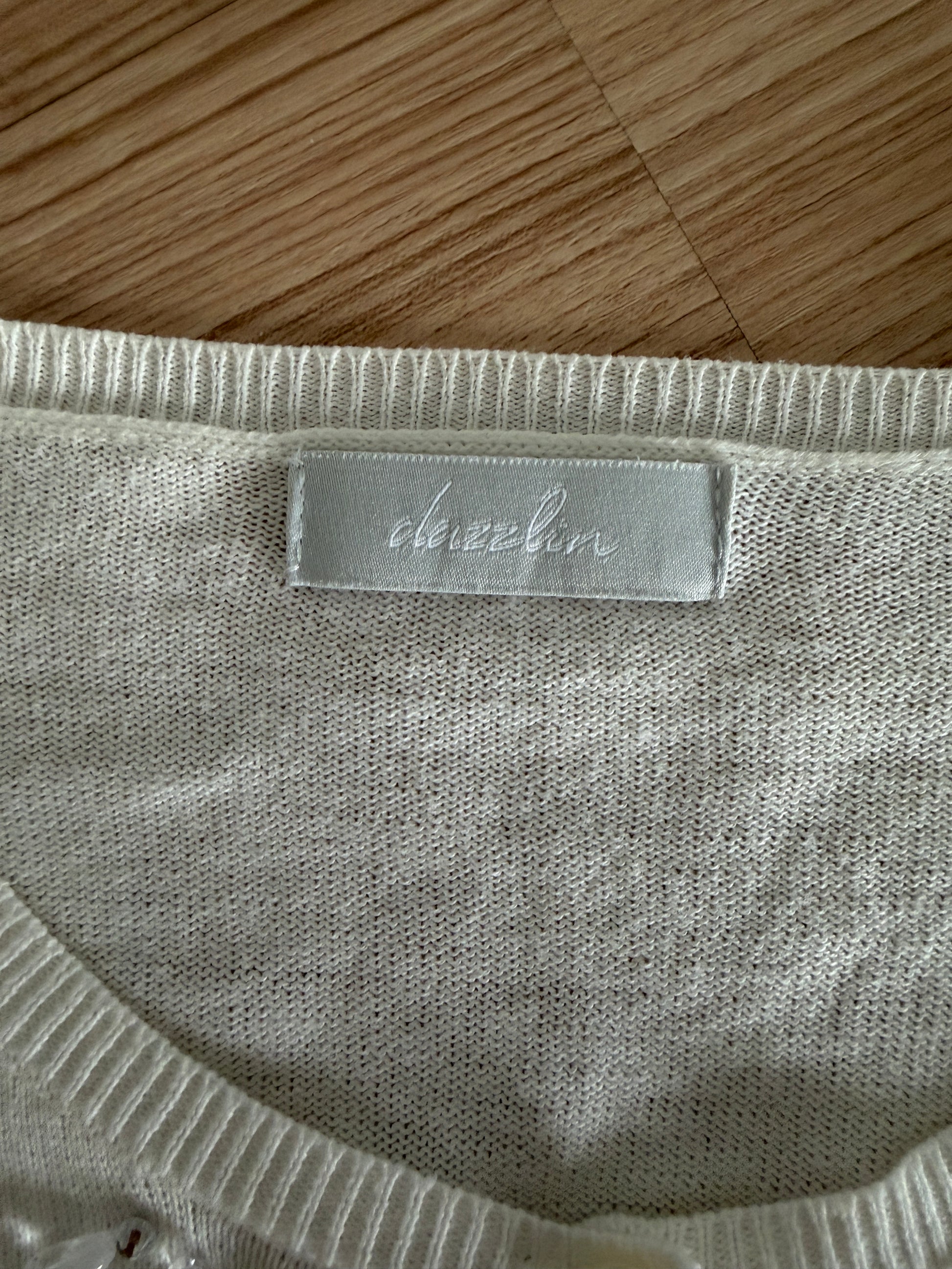 Close-up of a cream-colored knit garment with a silver-gray brand label reading 'dazzin' on a wooden surface.