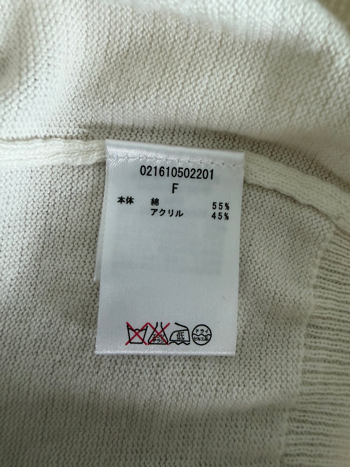 Close-up of a white knit garment label sewn onto a cream-colored sweater. The label shows product code 021610502201, size F, 55% acrylic, with care instruction symbols printed at the bottom.