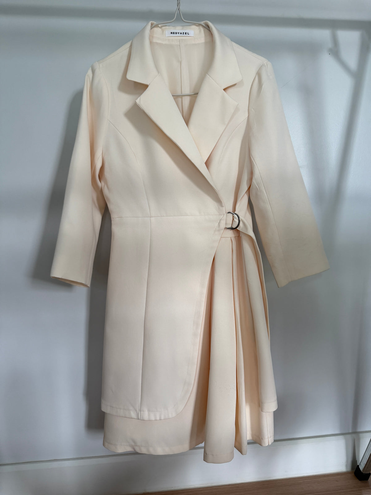 A light cream-colored wrap coat dress with three-quarter sleeves and a matching waist tie, hanging on a hanger against a white wall.