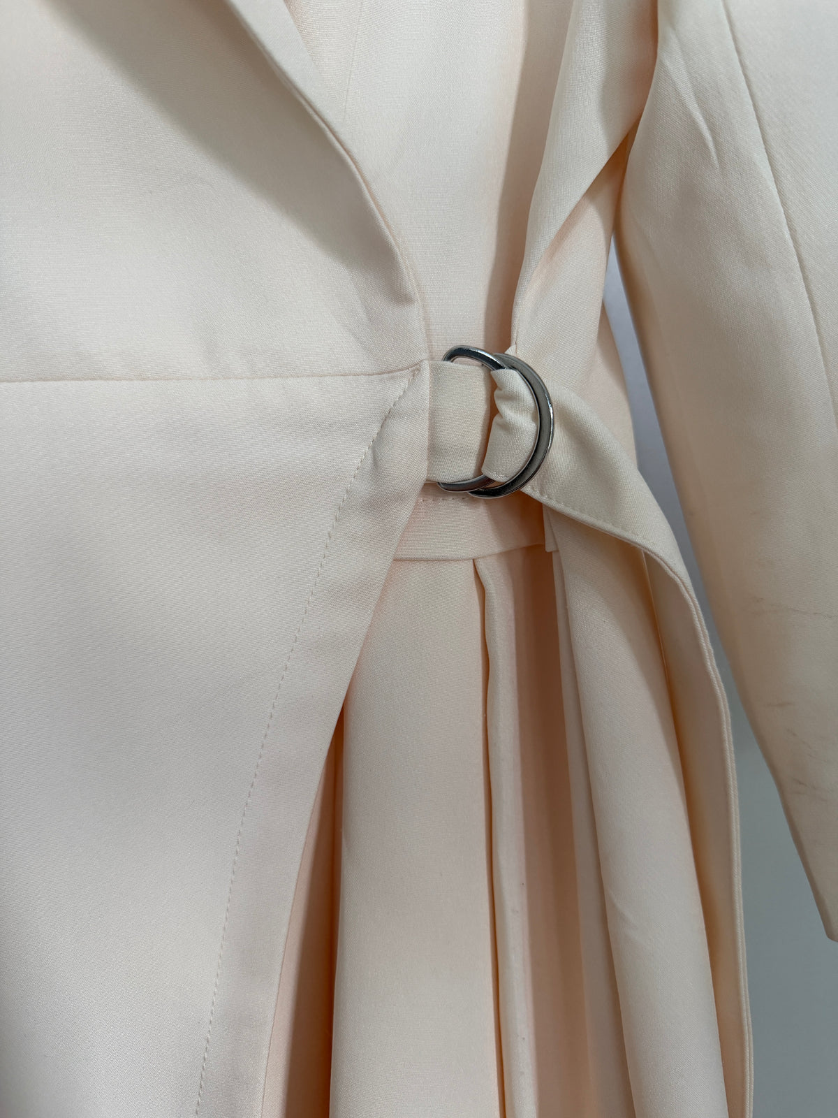 Close-up of a light cream or off-white tailored jacket or coat with a matching waist tie and a dark metal O-ring detail, shown against a neutral gray background.