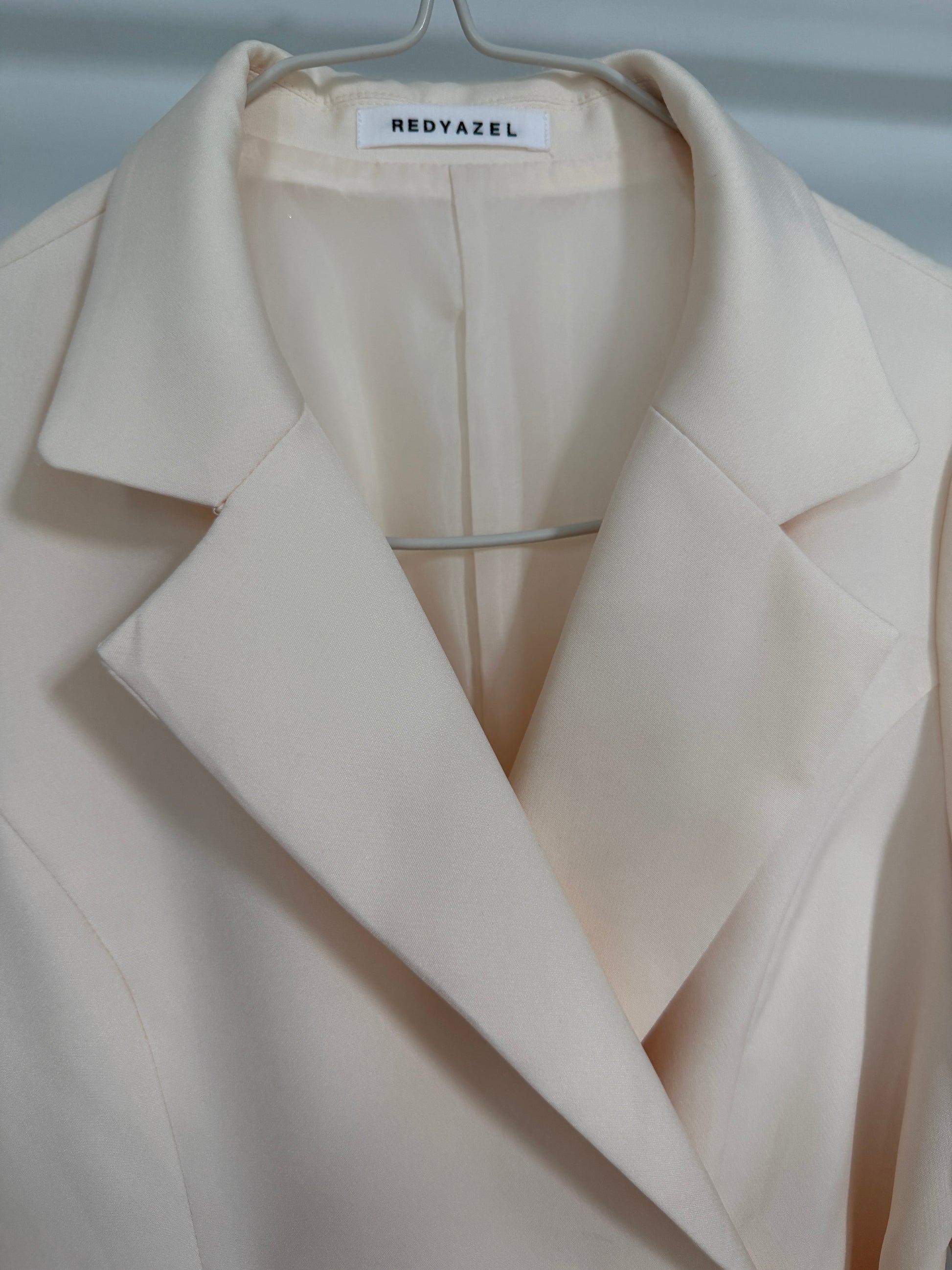 A close-up of a light cream or off-white blazer jacket with a structured collar, displayed on a hanger. The brand label 'REDYAZEL' is visible inside the collar.