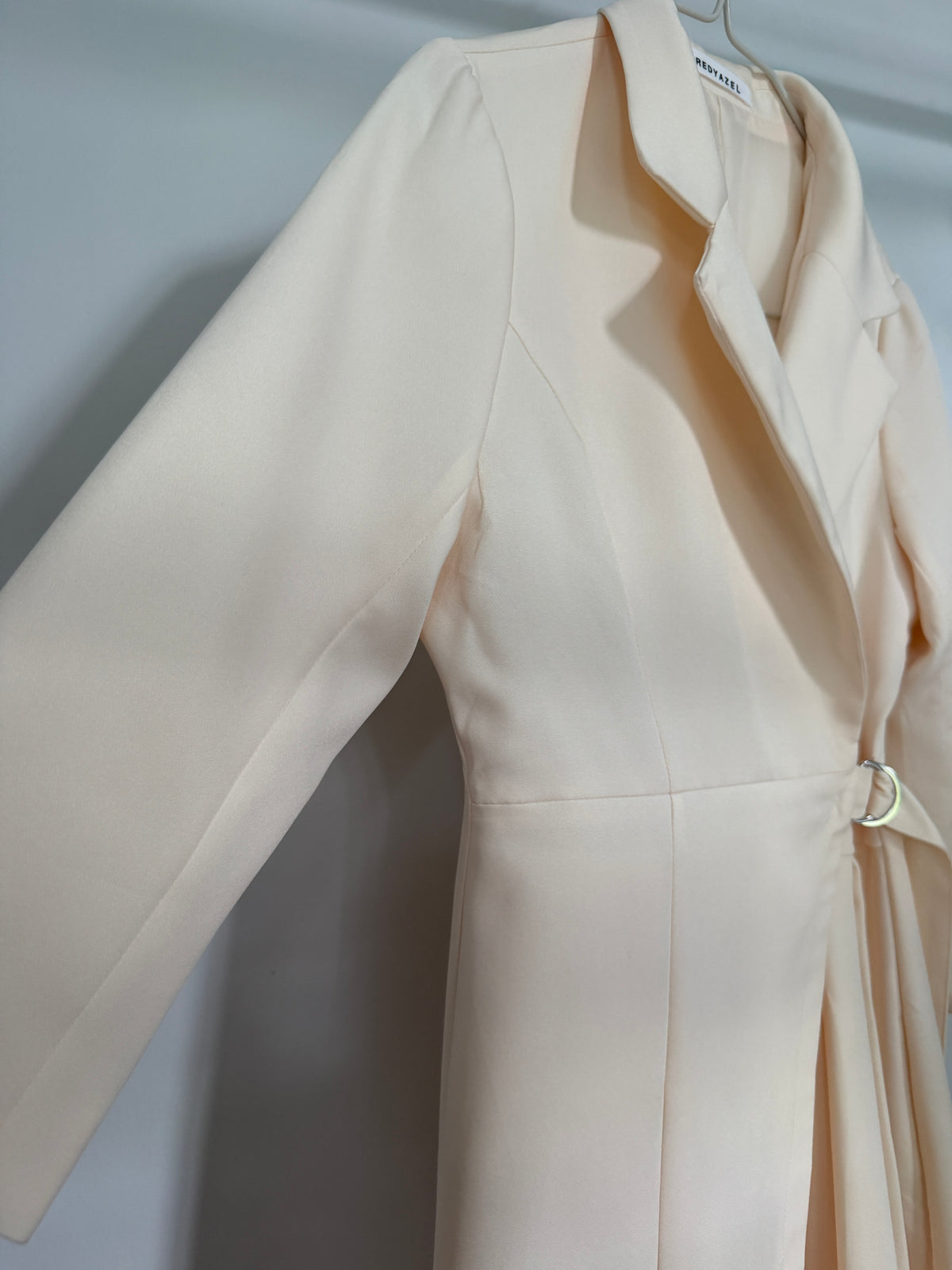 A light cream or pale peach wrap coat with a tie belt and a small gold ring accent, shown hanging against a neutral gray background.