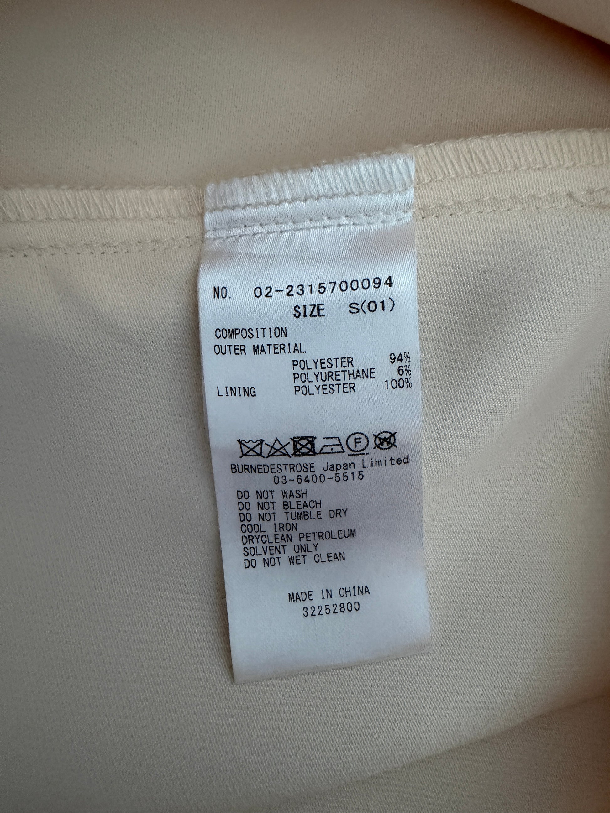 A close-up of a white fabric care label sewn onto the inside collar of a light beige garment. The label contains product information, material composition, and a MACH certification mark.