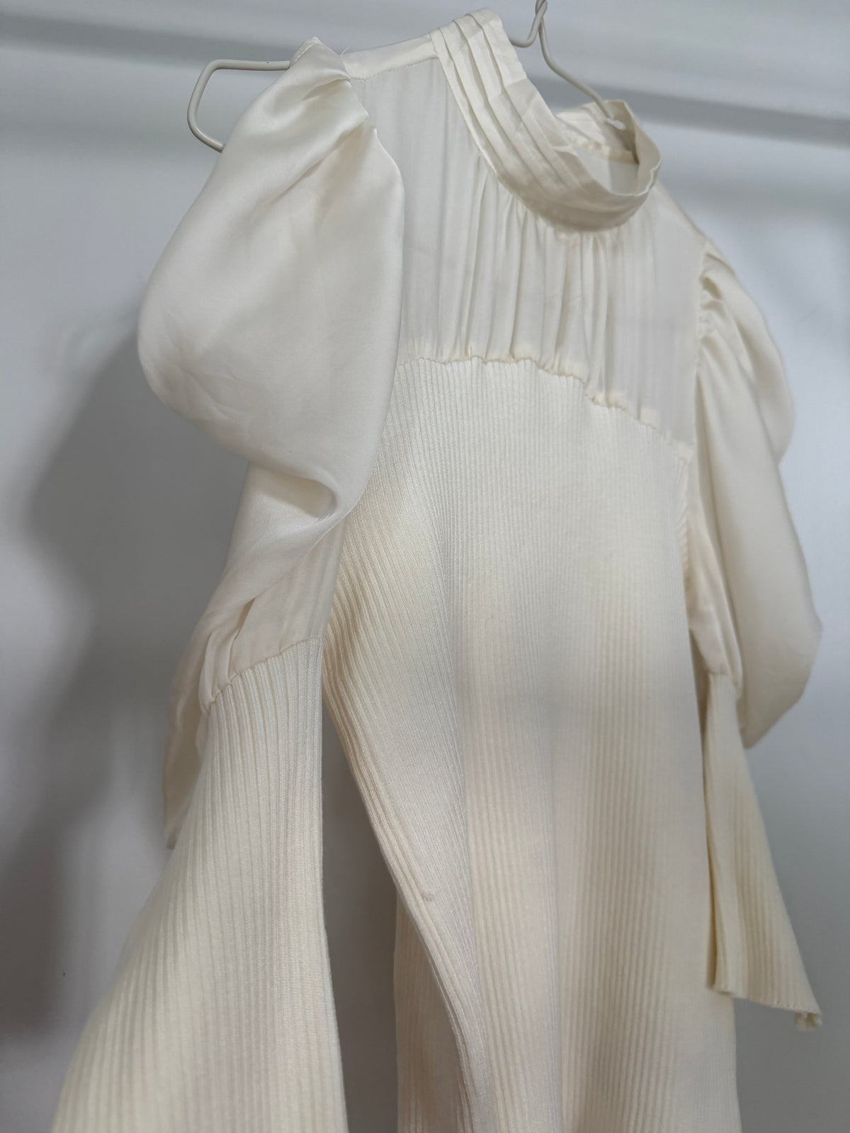 An off-white ribbed knit dress with dramatic puffed sleeves and a high gathered neckline, hanging on a metal hanger against a light gray background.