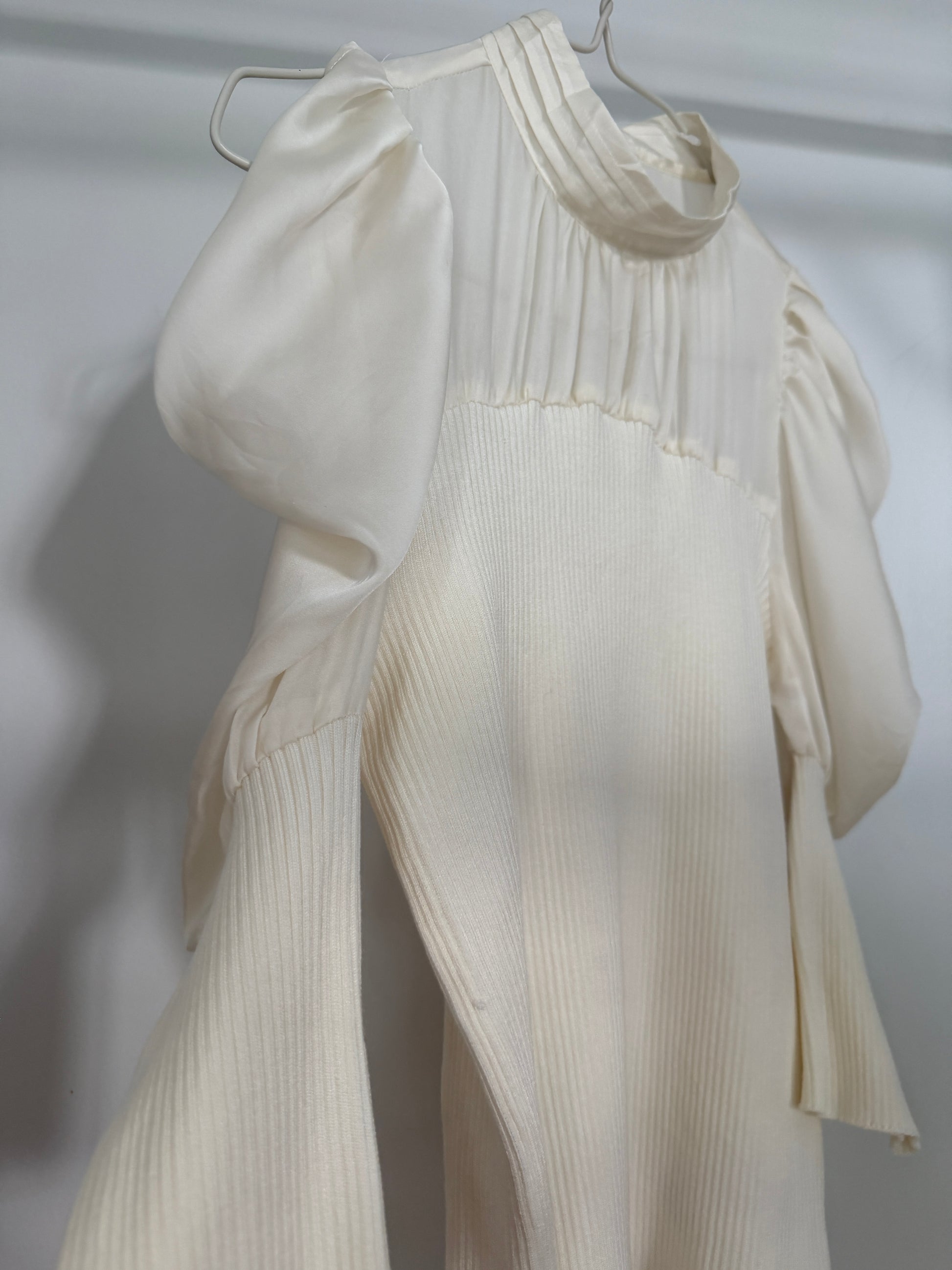 An off-white ribbed knit dress with dramatic puffed sleeves and a high gathered neckline, hanging on a metal hanger against a light gray background.