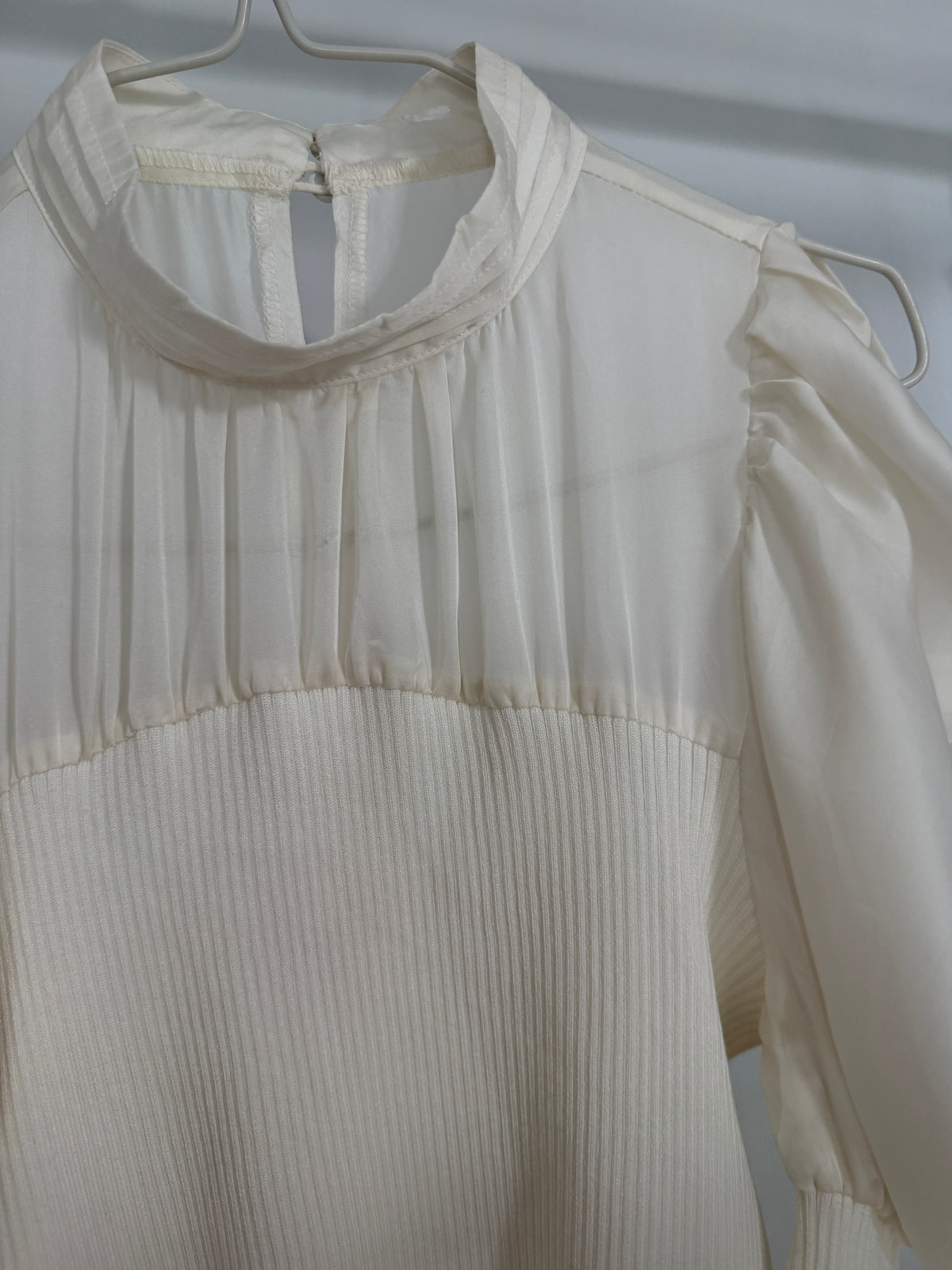 An off-white or cream-colored blouse with a high gathered mock neck, pleated bodice, and long sheer puff sleeves, hanging on a metal hanger against a light background.