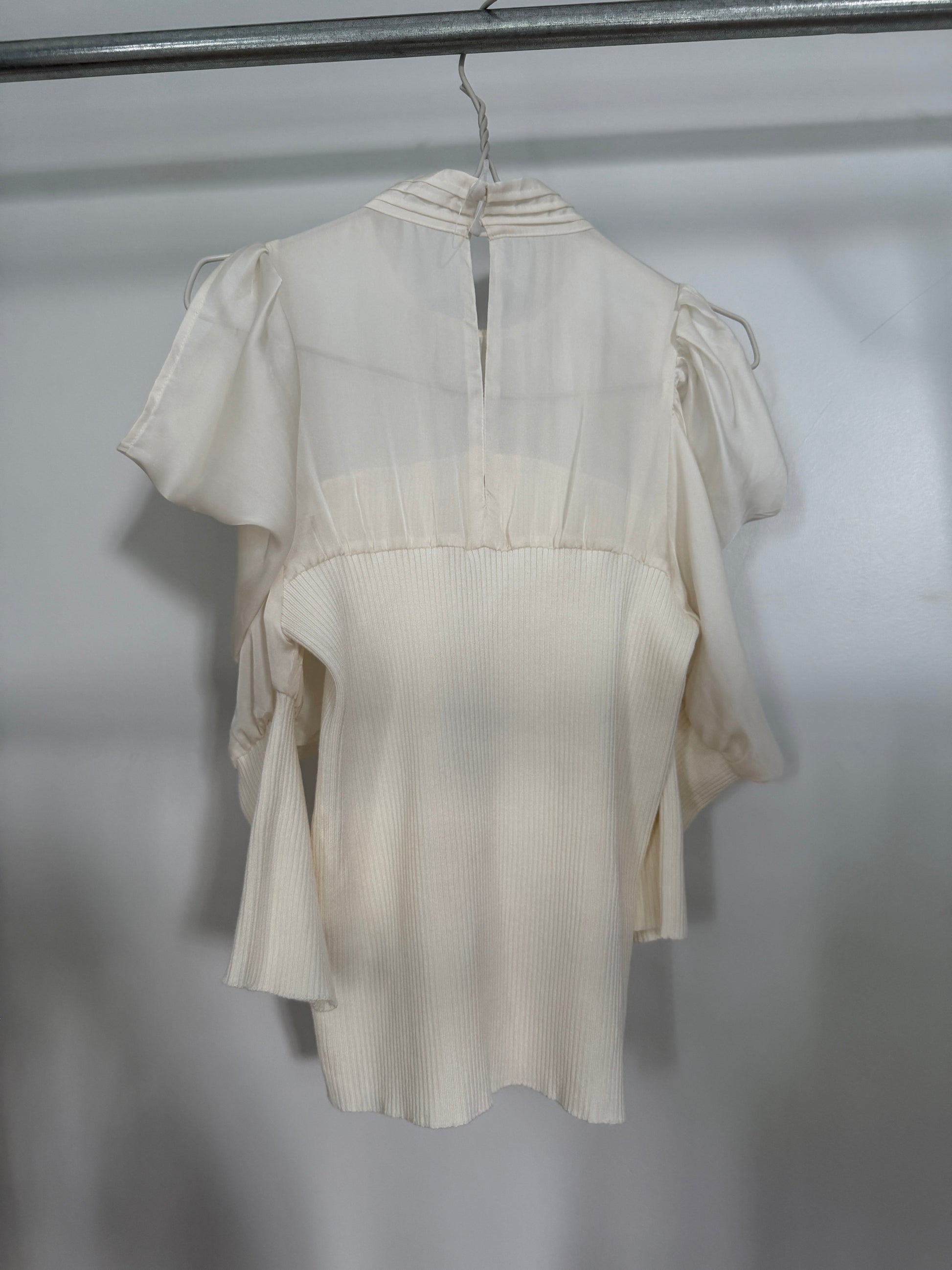 An off-white short-sleeve blouse with pleated details and a high neckline, hanging on a metal hanger against a plain light gray wall.