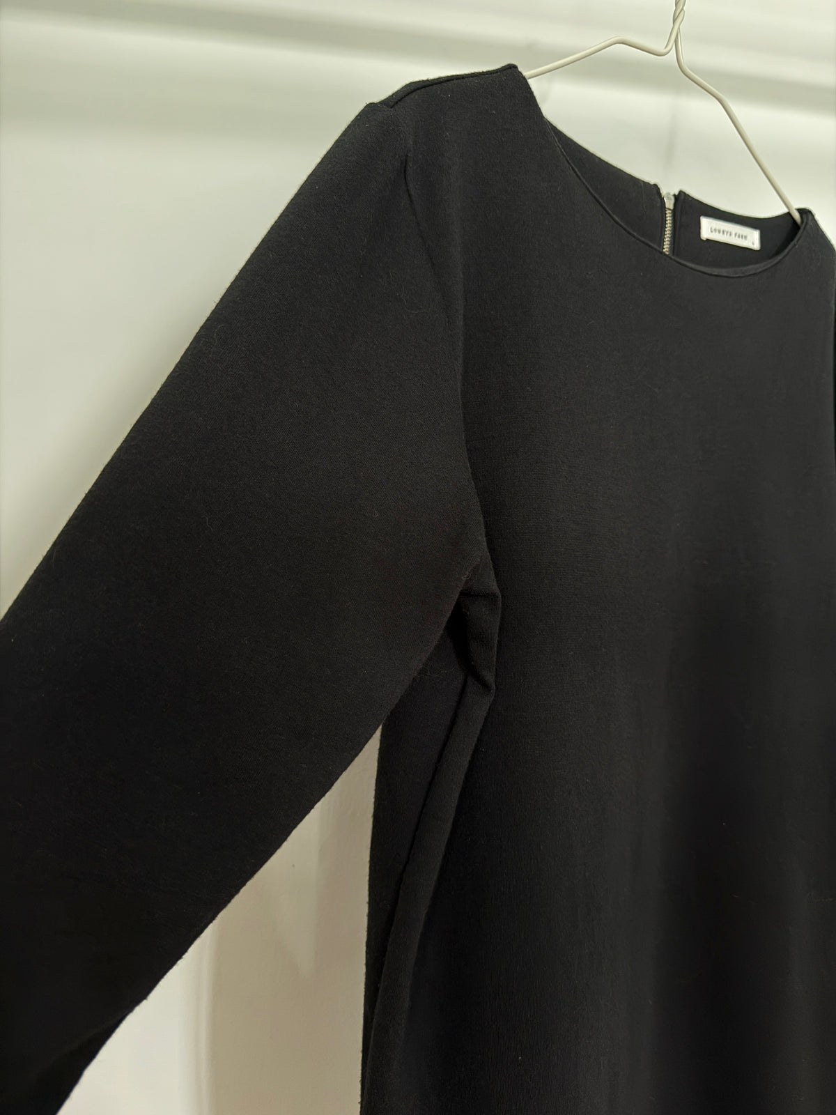 A black long-sleeve knit top hanging on a white hanger against a light gray fabric backdrop.