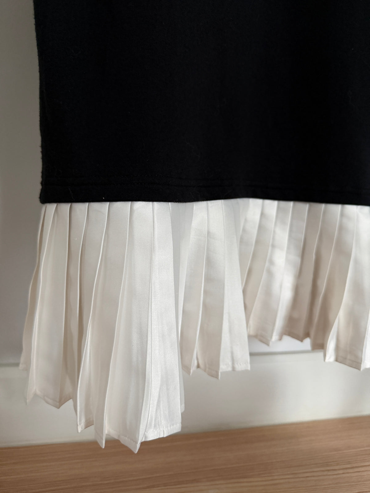 Close-up of a black garment with a layered white pleated hem, hanging against a light wall above a wooden surface.