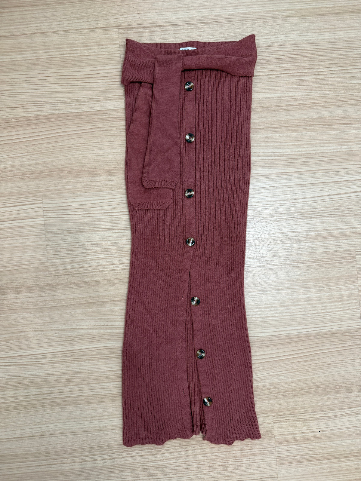 A ribbed knit skirt in a dusty rose or mauve color, laid flat on a light wood-grain floor. The skirt features a tie-waist belt, a front button closure, and a flared silhouette.