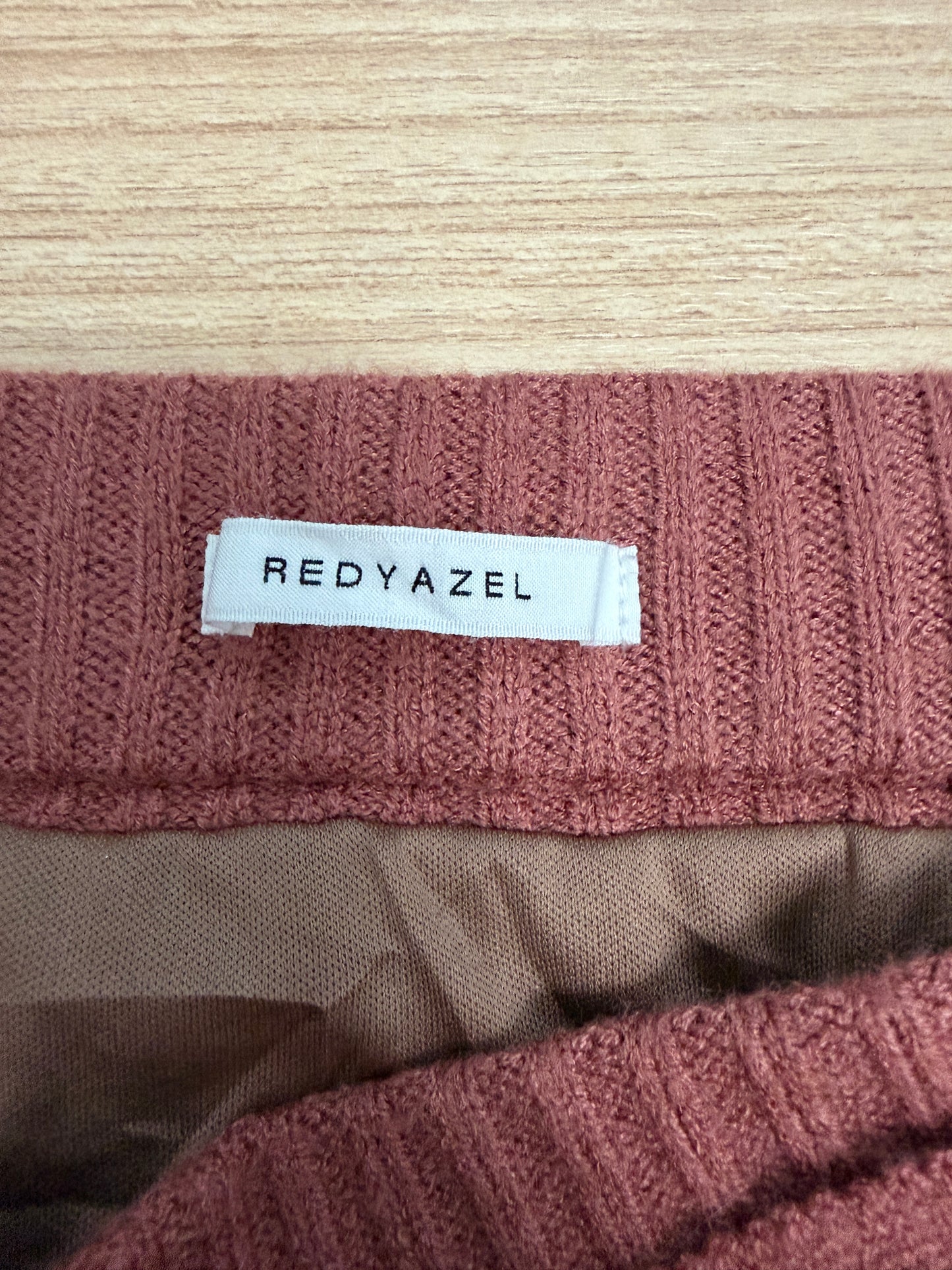Close-up of a ribbed knit garment in dusty rose pink with a white sewn-in label reading REDYAZEL, placed on a light wood surface.