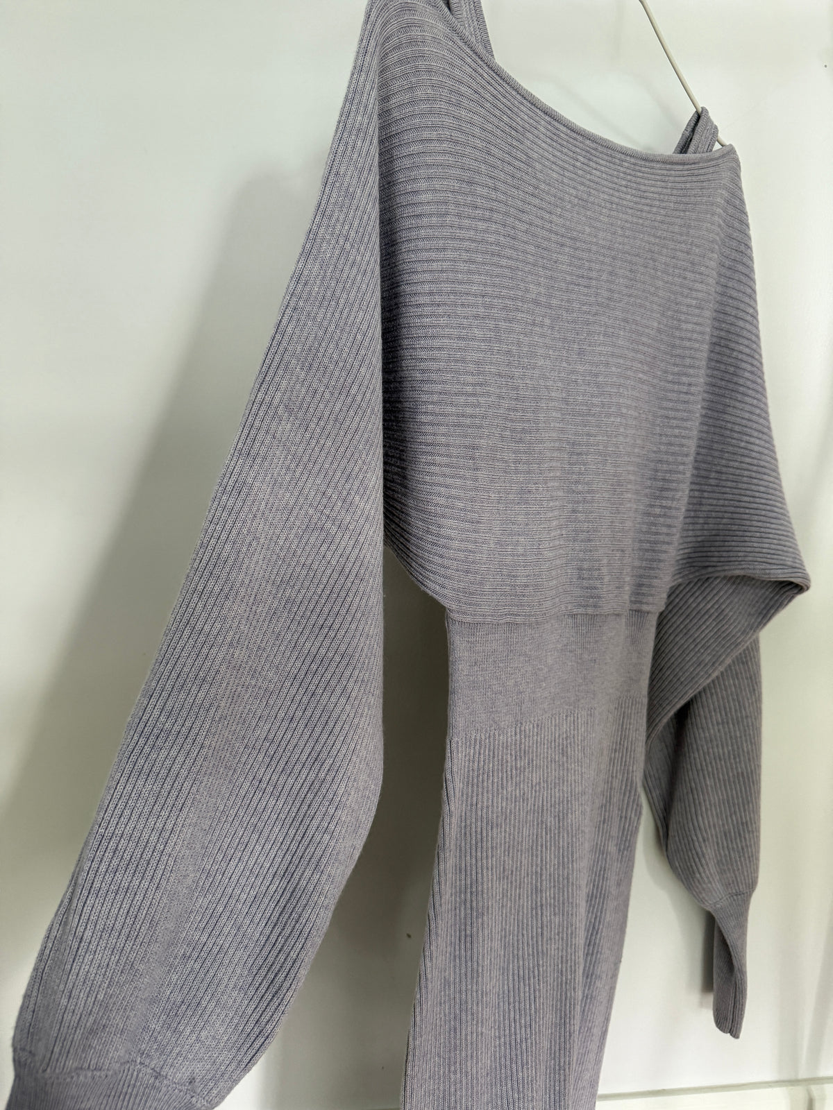 A light gray ribbed knit long-sleeve top with an asymmetrical draped neckline and layered front, hanging against a plain light background.