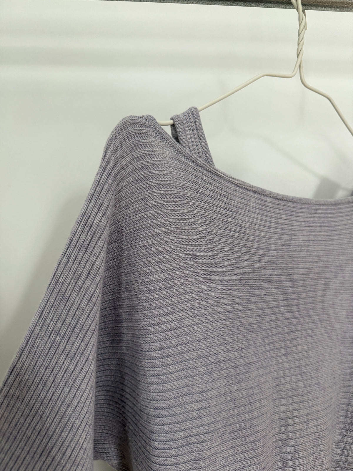 A close-up of a ribbed knit top in a muted grayish purple/lavender color, hanging on a white wire hanger against a plain light background.