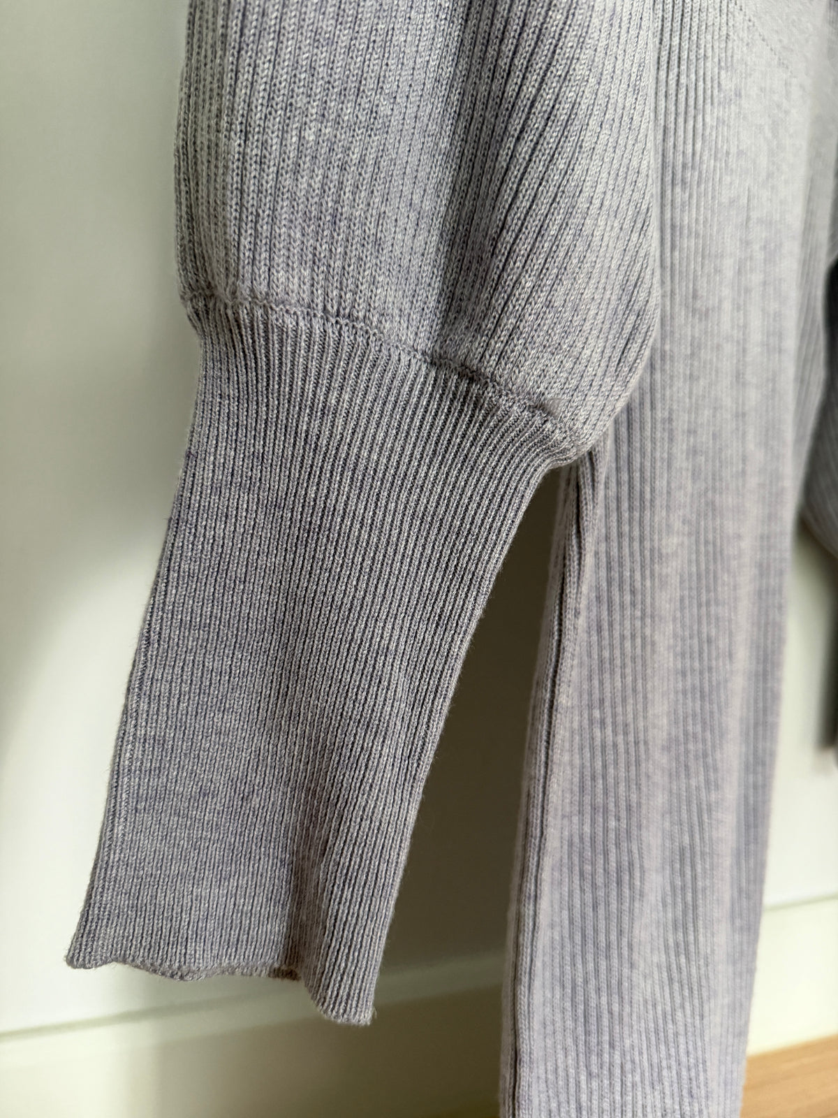 Close-up of a light gray ribbed knit garment, showing the sleeve cuff and vertical rib texture against an off-white wall.