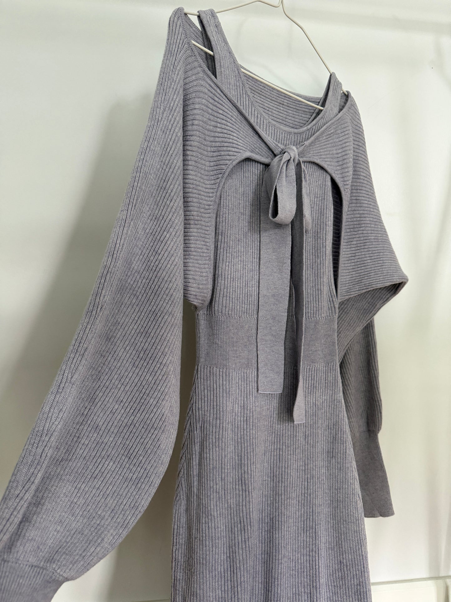 A gray ribbed knit long-sleeve dress with a tie-neck detail, hanging on a metal hanger against a white fabric backdrop.