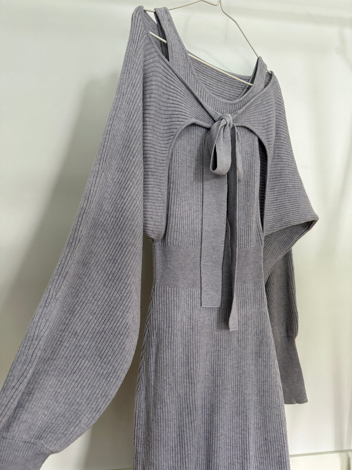 A gray ribbed knit long-sleeve dress with a tie-neck detail, hanging on a metal hanger against a white fabric backdrop.
