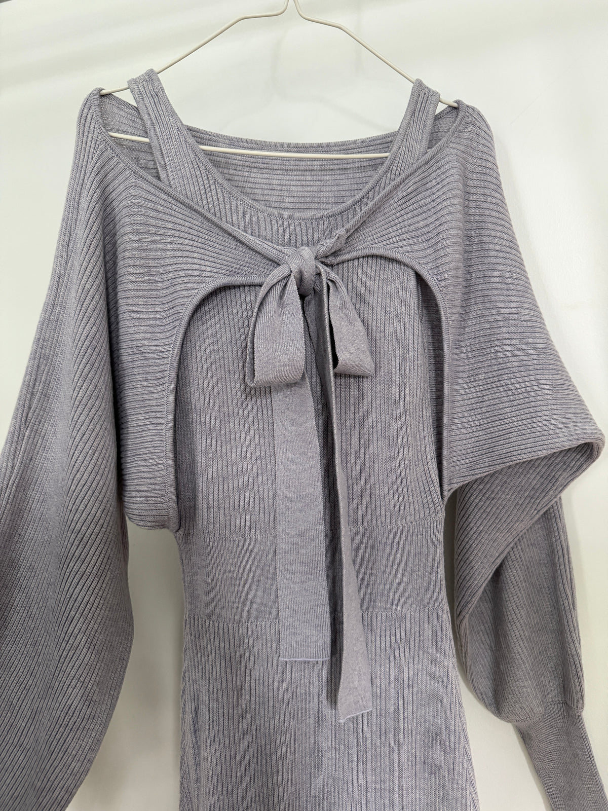 A light gray ribbed knit long-sleeve top with a wide scoop neckline, shoulder cutouts, and a front tie detail, hanging on a white hanger against a white background.