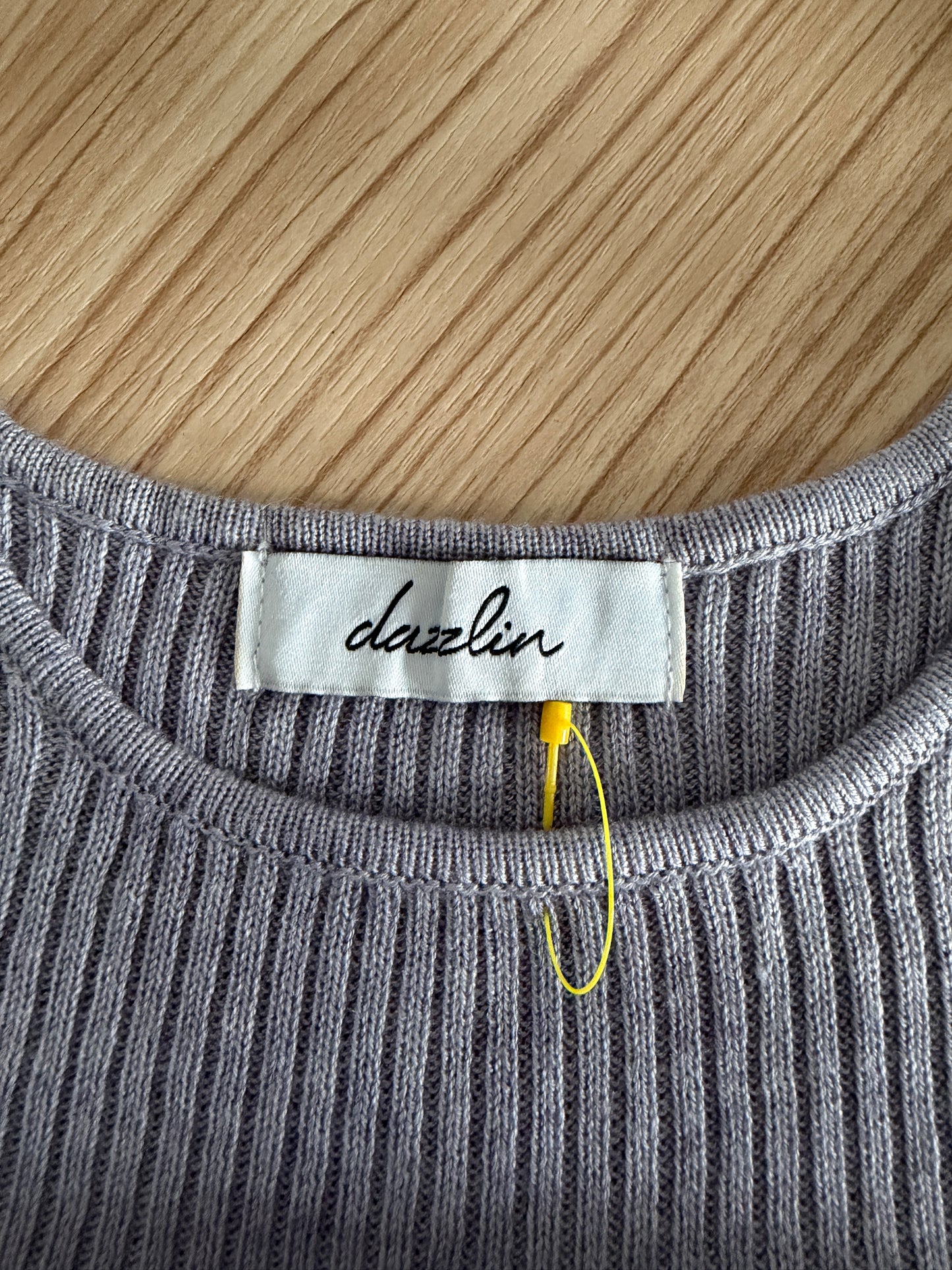 Close-up of a gray ribbed knit crewneck sweater showing the white 'dazlin' brand label and a yellow hangtag, laid flat on a light wood background.