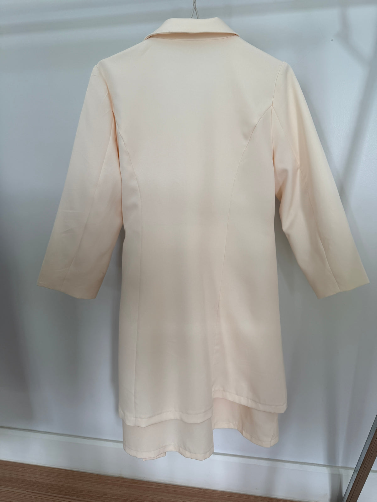 A light cream or pale peach-colored long-sleeve shirt or tunic hanging on a hanger against a white wall.