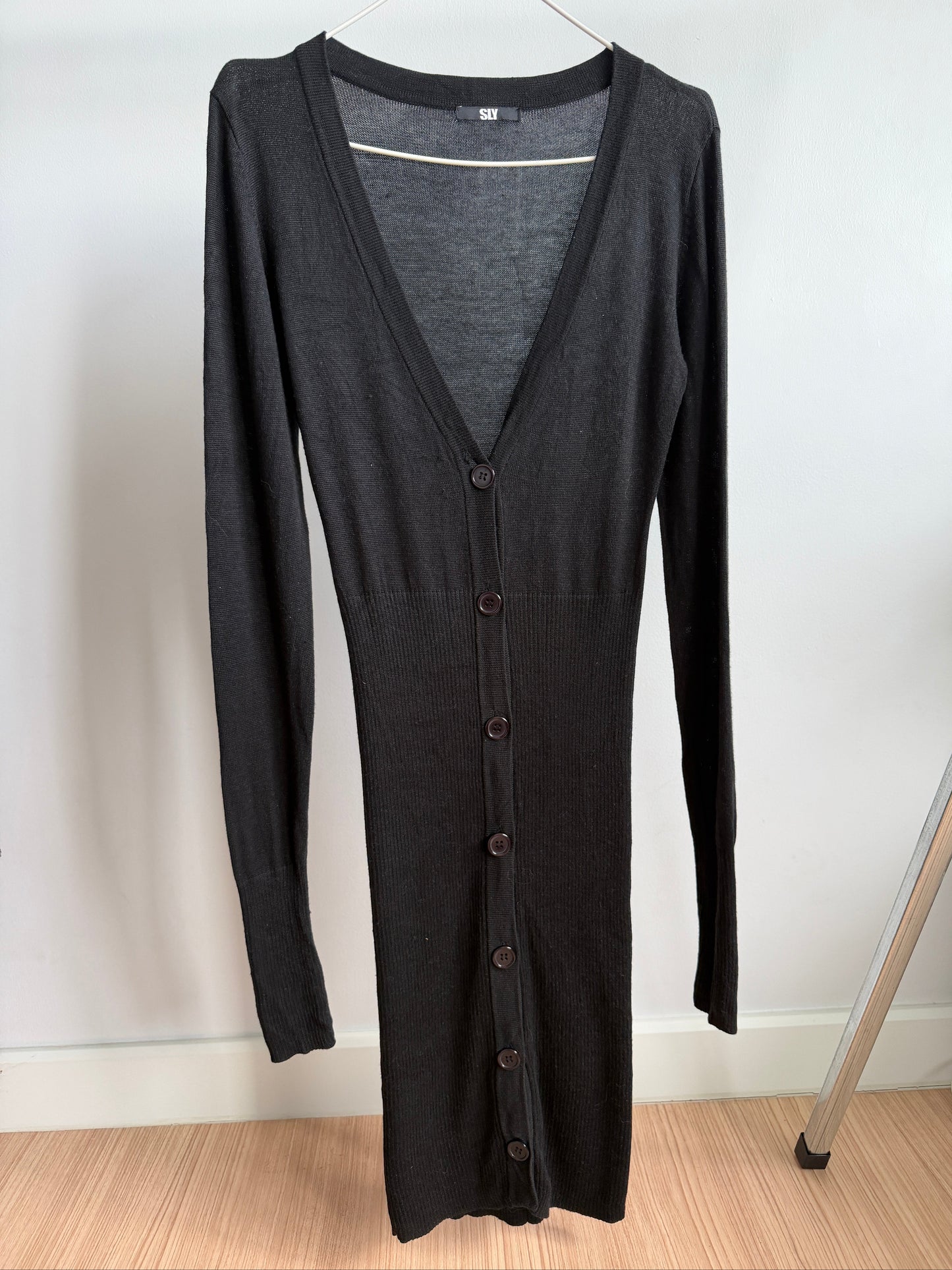 A dark charcoal gray long-sleeve button-front cardigan dress hanging on a hanger against a plain white wall, with a light wood floor visible at the bottom.