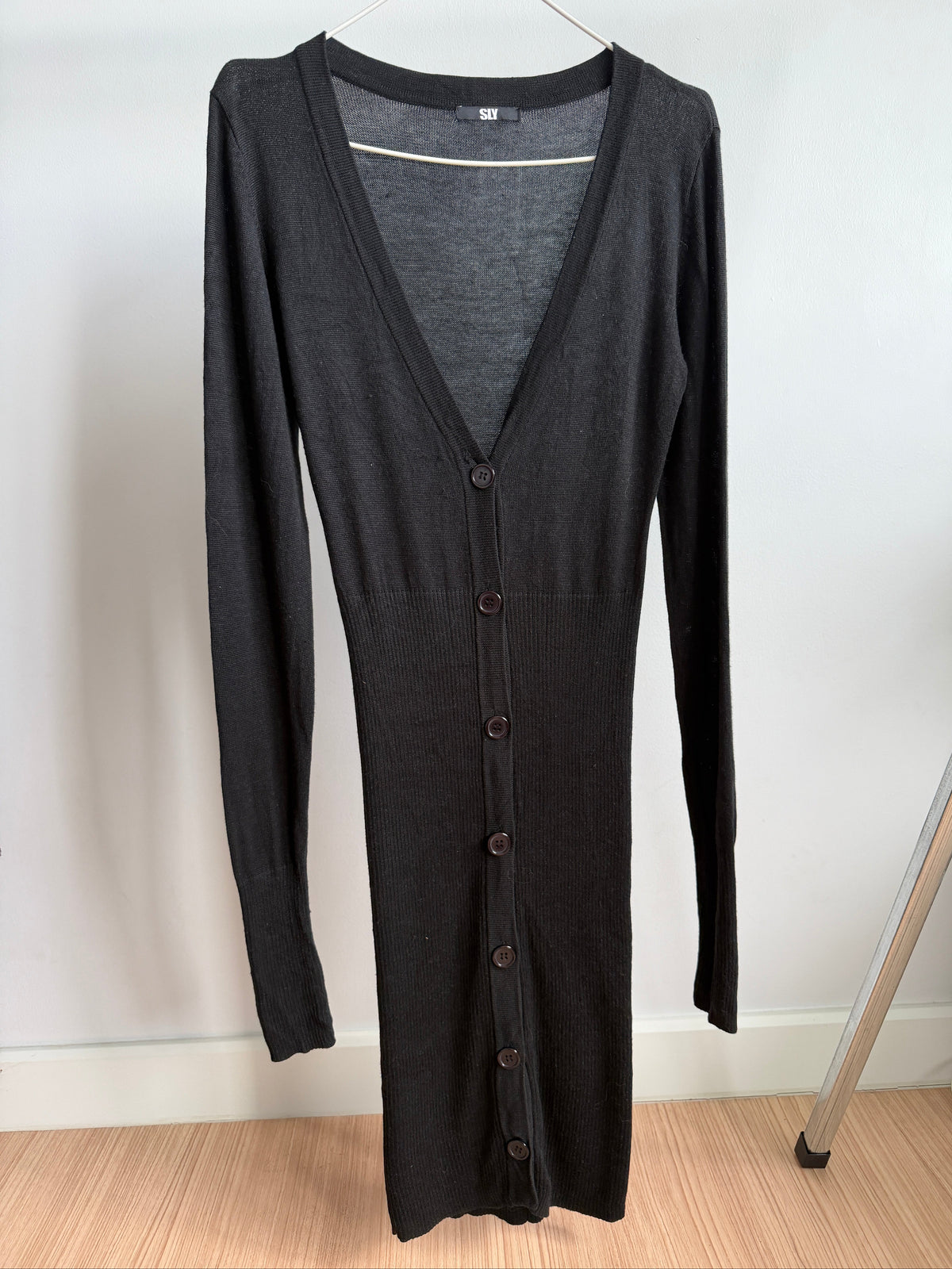 A dark charcoal gray long-sleeve button-front cardigan dress hanging on a hanger against a plain white wall, with a light wood floor visible at the bottom.