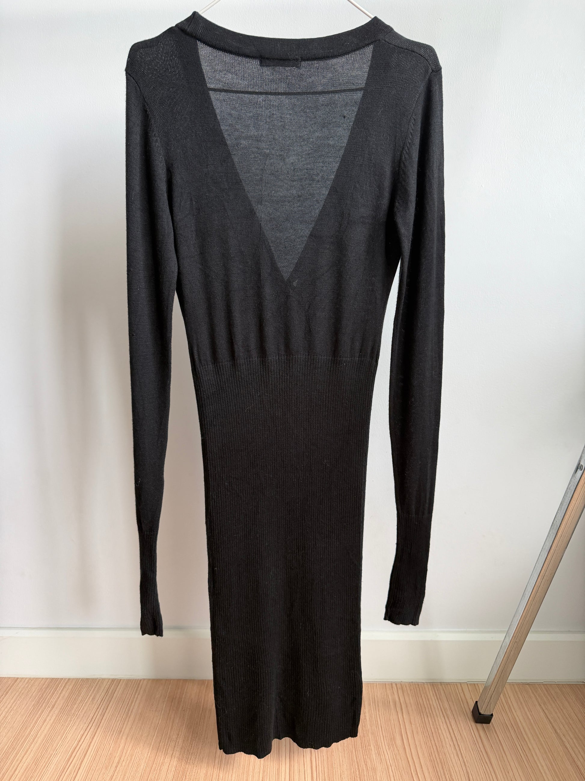A long-sleeve black knit dress with a deep V-neckline, hanging on a hanger against a white wall with a light wood floor.