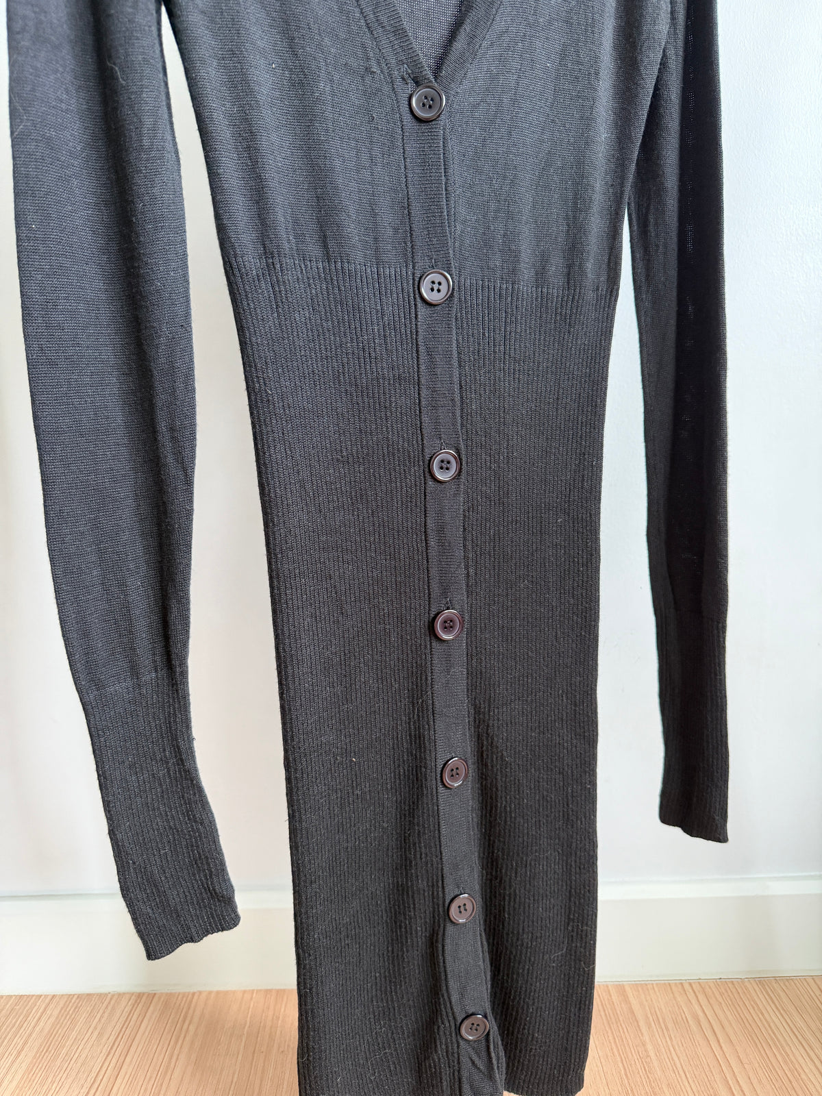 A dark gray ribbed knit cardigan dress with a V-neck and front button closure, shown hanging against a plain white wall with a light wood floor visible at the bottom.