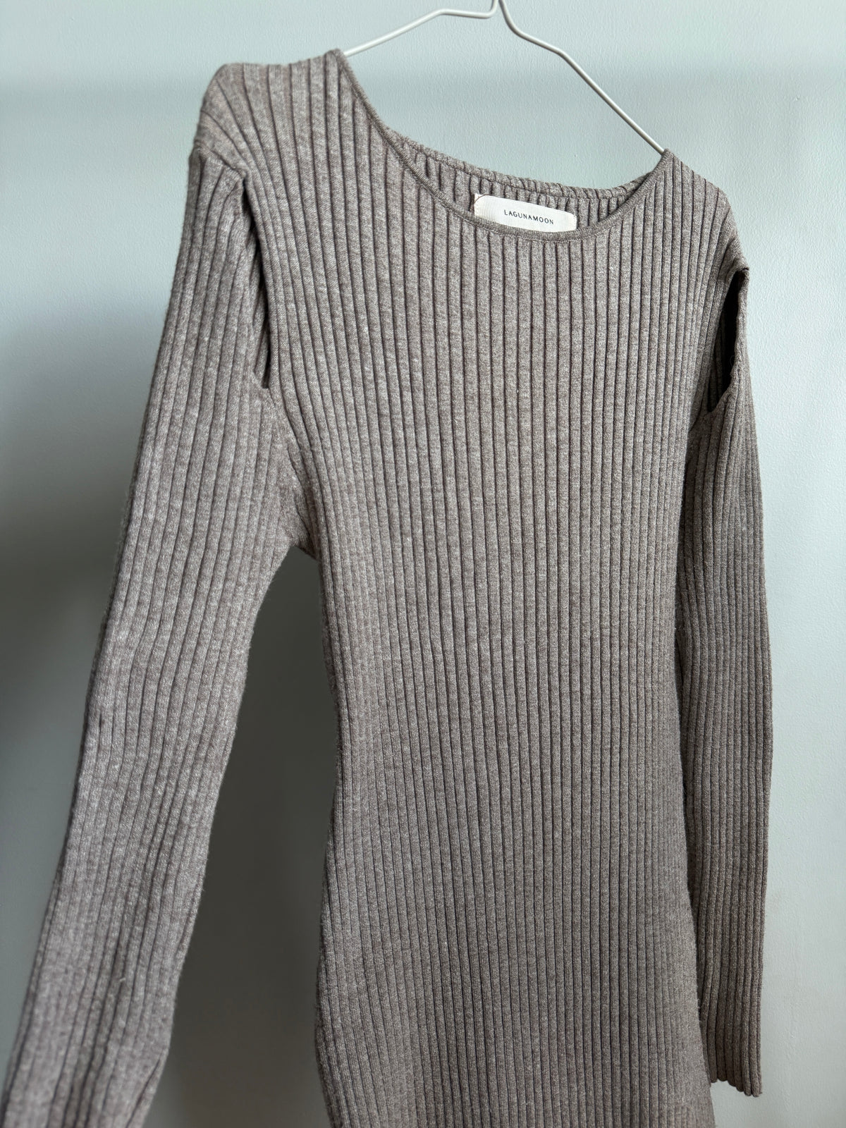A taupe ribbed knit long-sleeve sweater hanging on a metal hanger against a plain light gray wall.