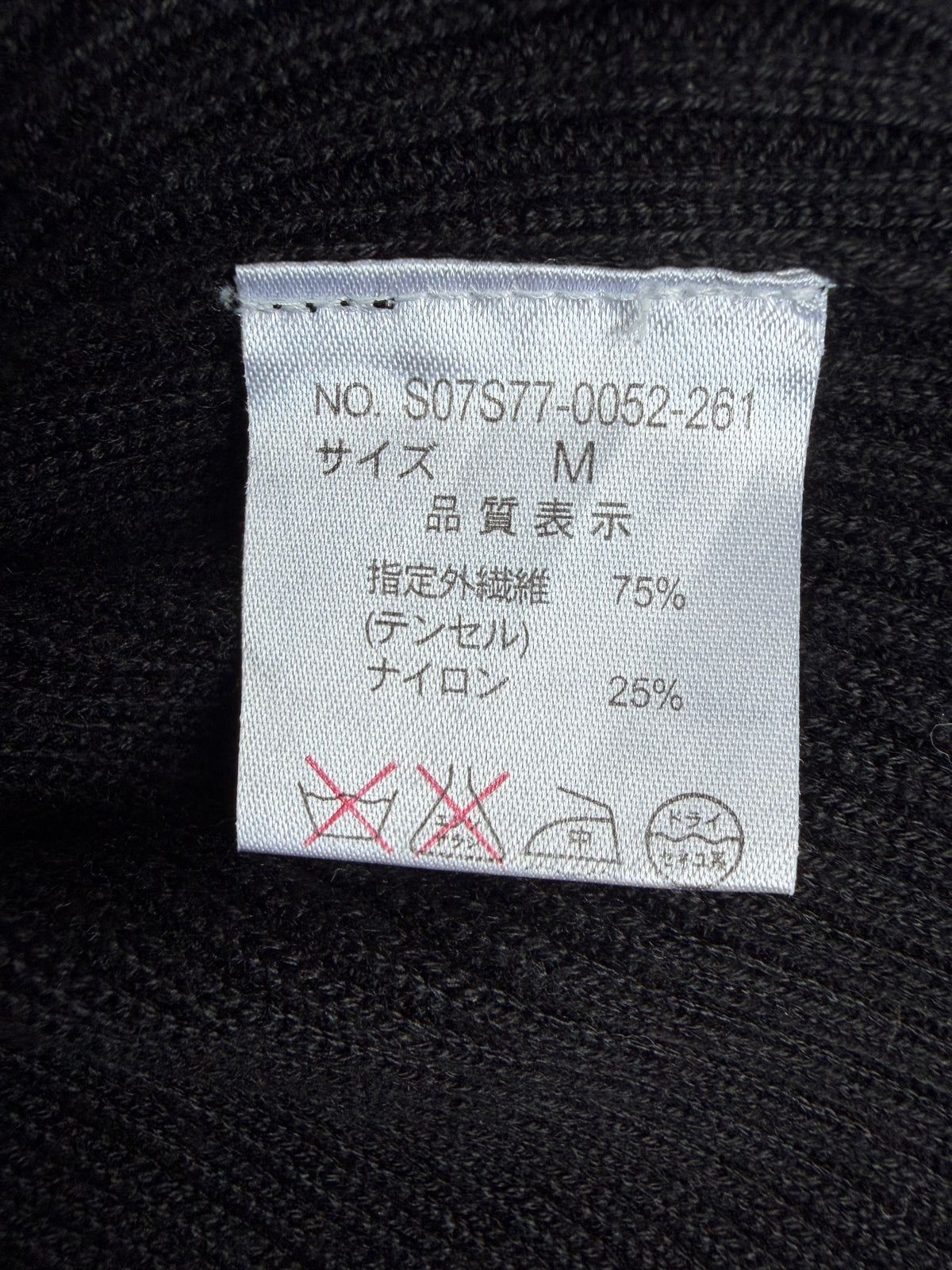 A close-up of a white fabric care label sewn onto a black knit garment, showing Japanese text with size M and material percentages.