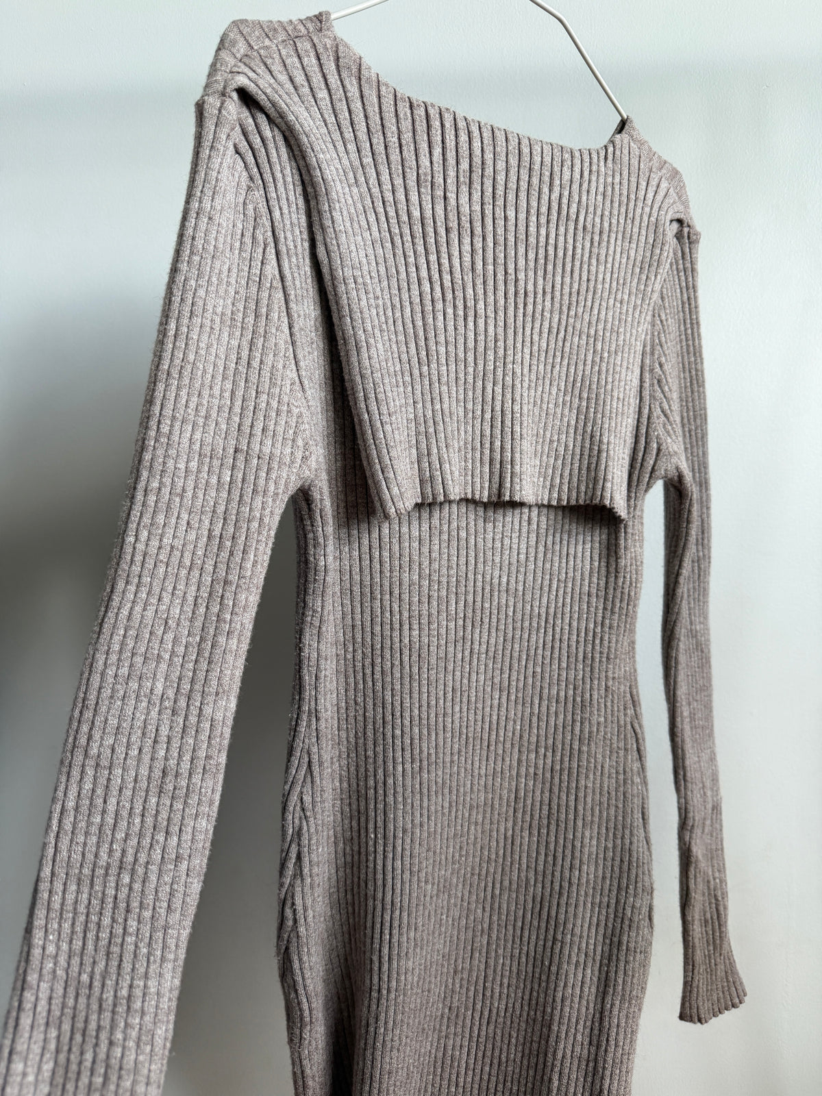 A ribbed knit long-sleeve dress in a taupe-gray color, hanging on a metal hanger against a plain light gray wall.