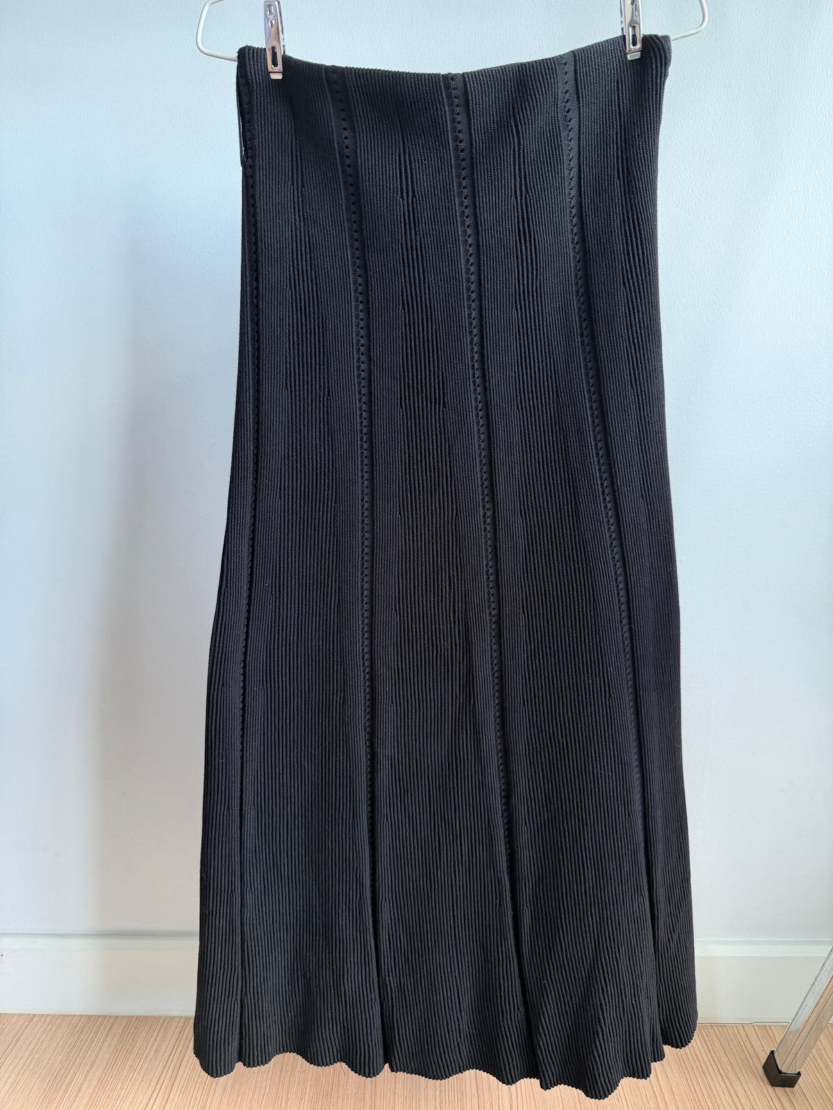 A black pleated midi skirt hanging from a metal hanger against a light gray wall and wood floor.