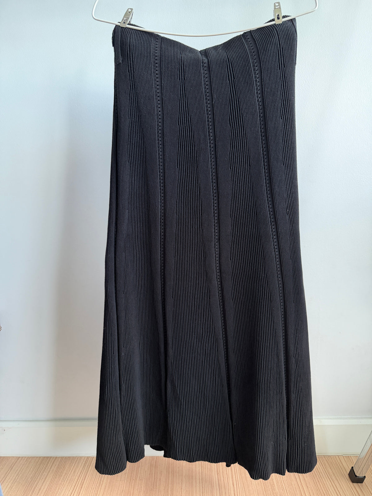 A dark charcoal gray pleated midi skirt hanging on a metal hanger against a plain white wall.