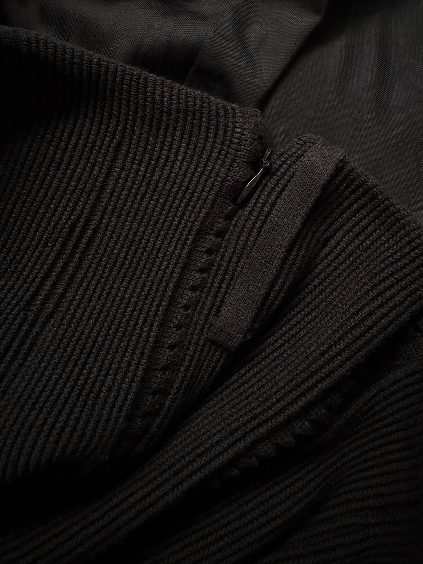 Close-up detail of a black ribbed knit garment, showing the texture of the ribbed fabric and a small metal zipper pull.
