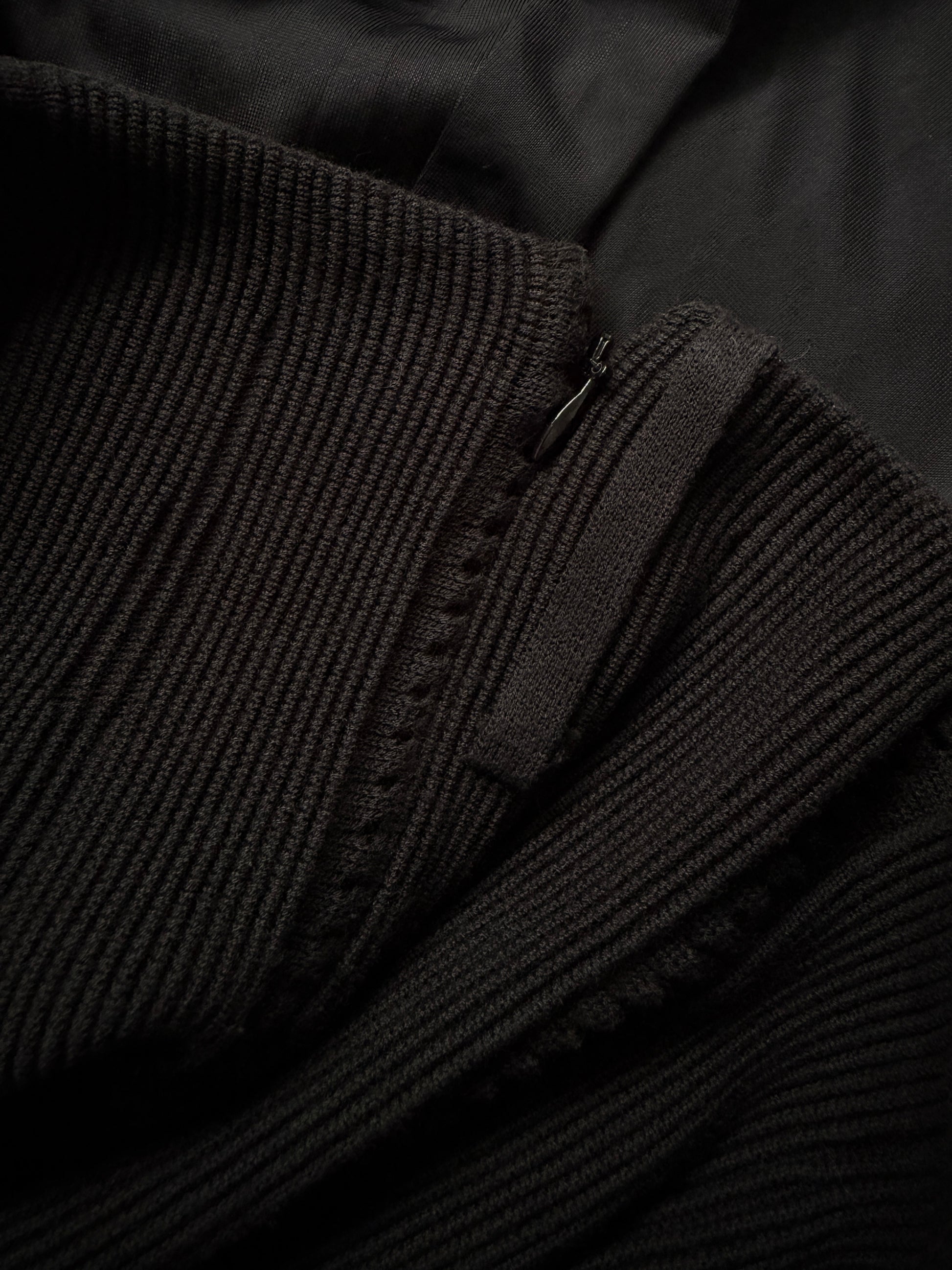 Close-up detail of a black ribbed knit garment, showing the texture of the ribbed fabric and a small metal zipper pull.
