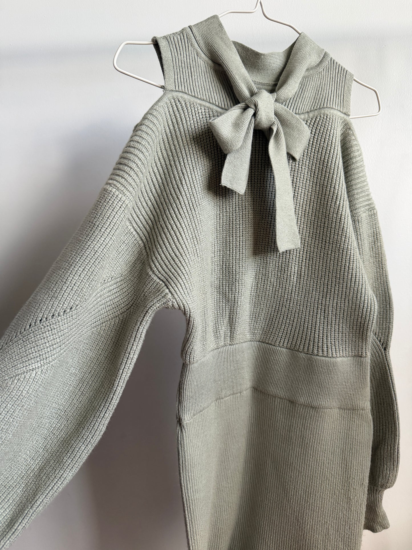A light sage green ribbed knit sweater with a matching fabric bow at the collar, hanging on a white wire hanger against a plain light gray wall.
