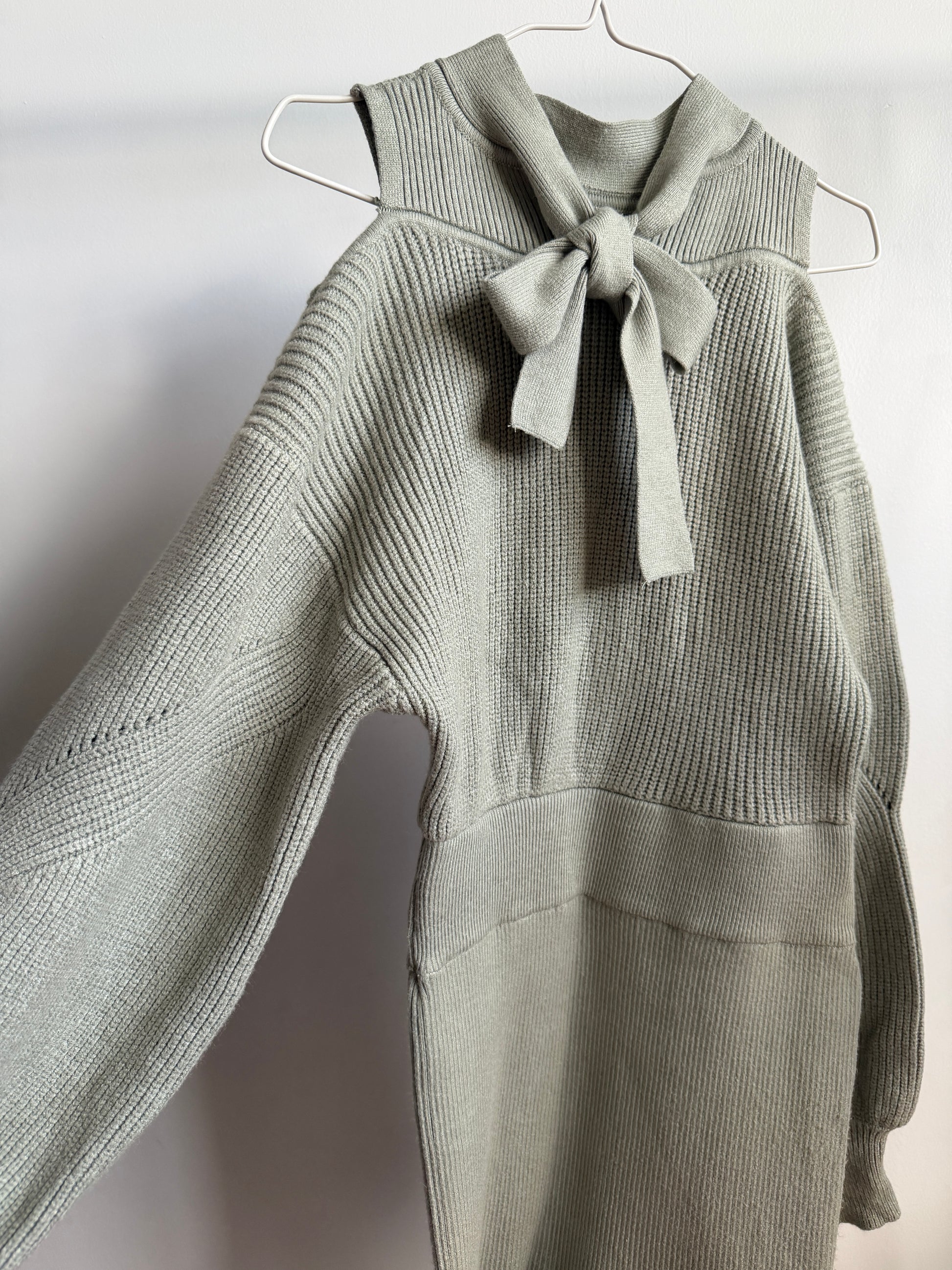 A light sage green ribbed knit sweater with a matching fabric bow at the collar, hanging on a white wire hanger against a plain light gray wall.