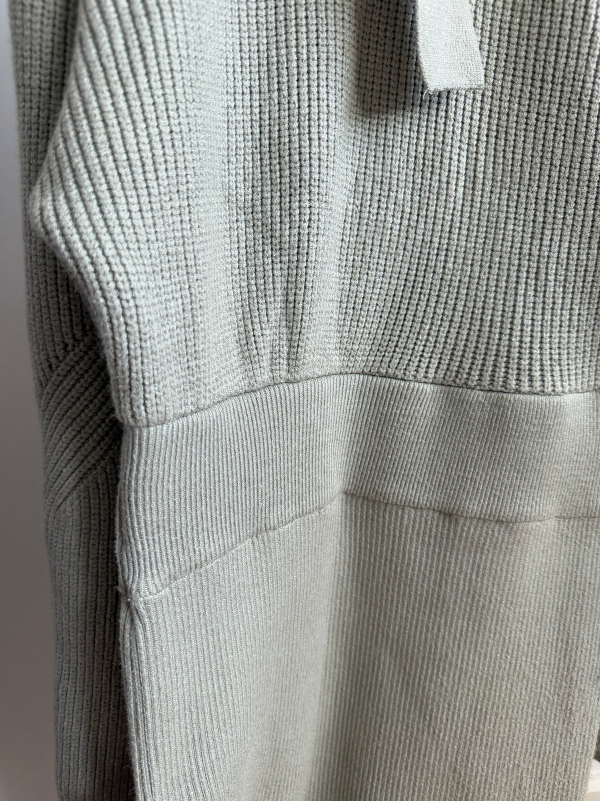 Close-up of a ribbed knit sweater in a light sage green color, showing the texture and a portion of the sleeve.
