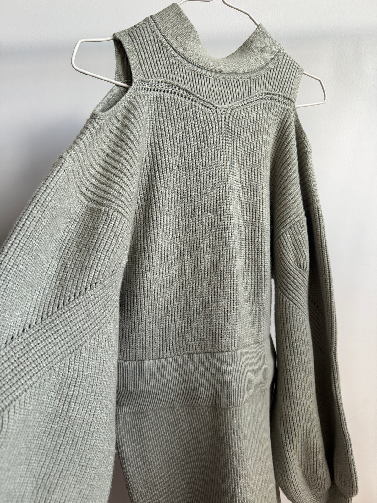 A light sage green ribbed knit sweater with a mock neck and asymmetrical shoulder cutouts, hanging on a white wire hanger against a plain white wall.
