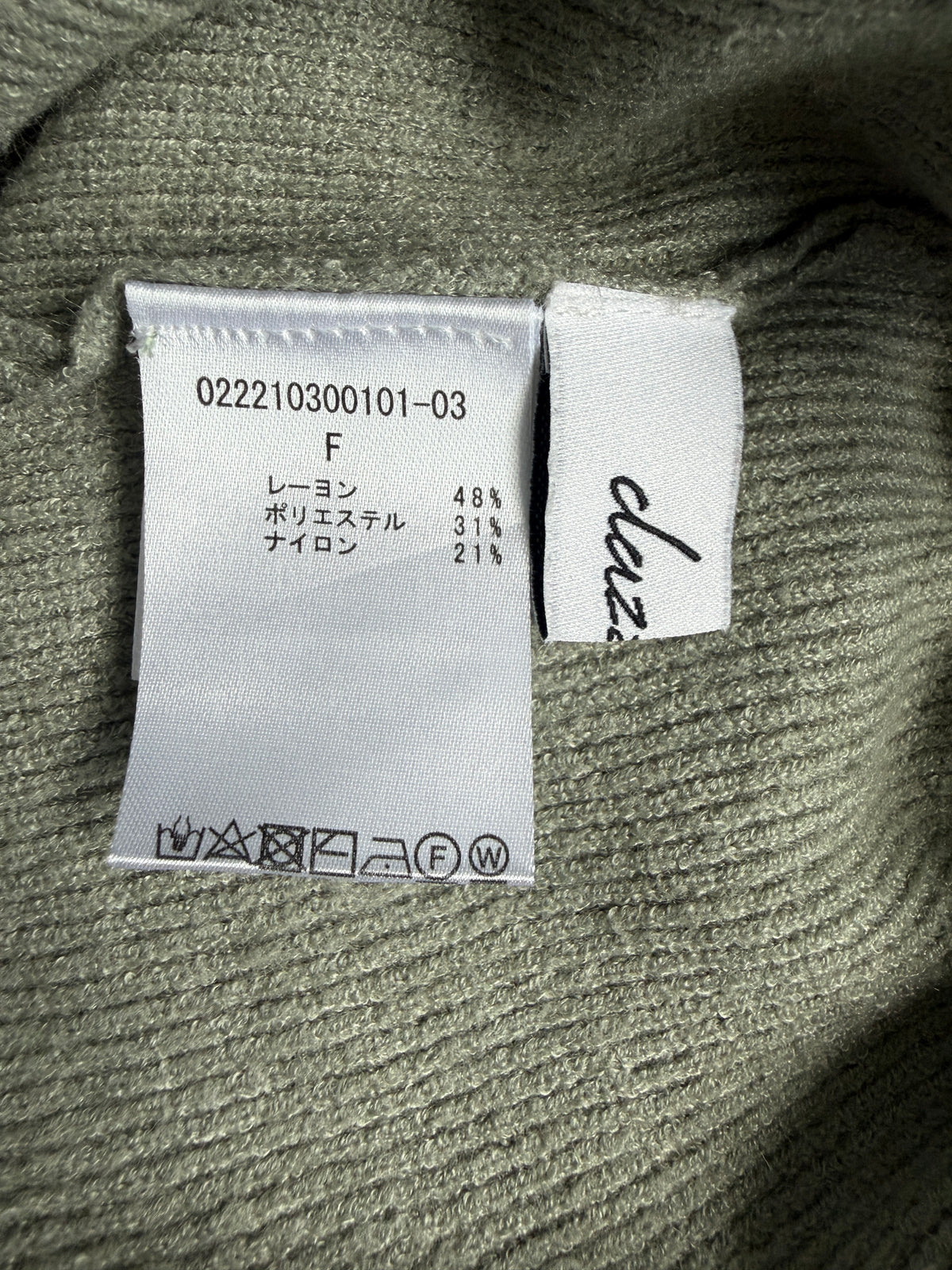 Close-up of a ribbed knit garment label sewn onto an olive green sweater. The white care label shows product code 022210300101-03, composition with 48% rayon, 31% nylon, and 21% other fibers, and a brand tag with the script name 'clazee'.