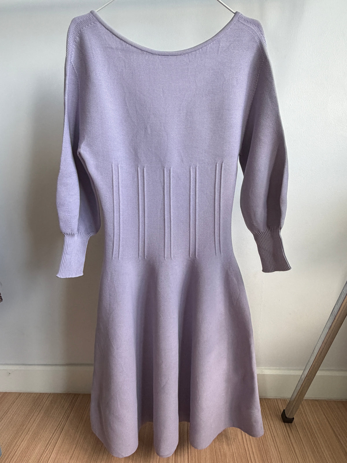 A light lavender knit dress with three-quarter sleeves and a flared skirt, hanging on a metal hanger against an off-white wall.