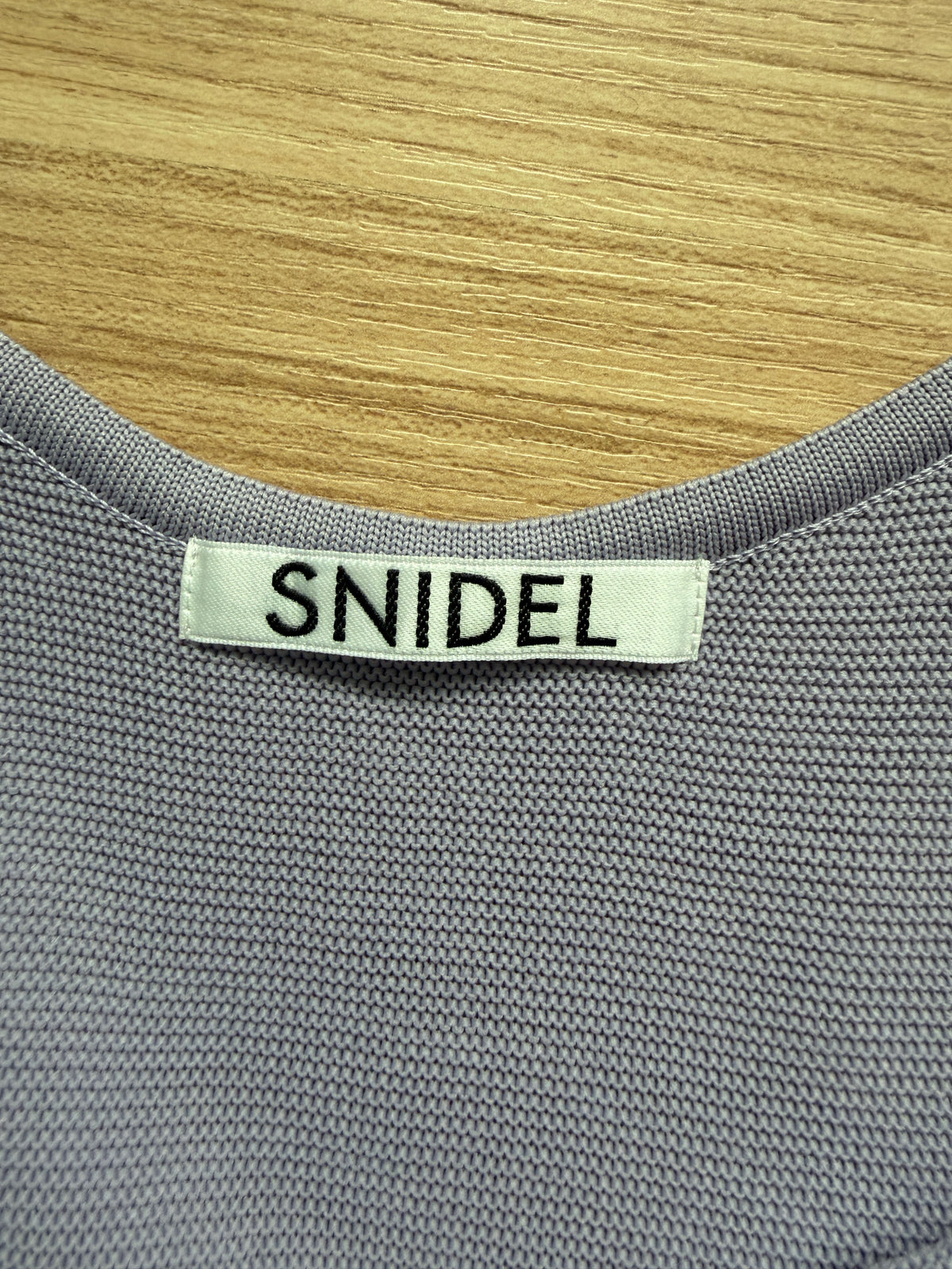 Close-up of a gray ribbed knit garment with a white fabric neck label reading 'SNIDEL' in black text, placed against a light wood background.