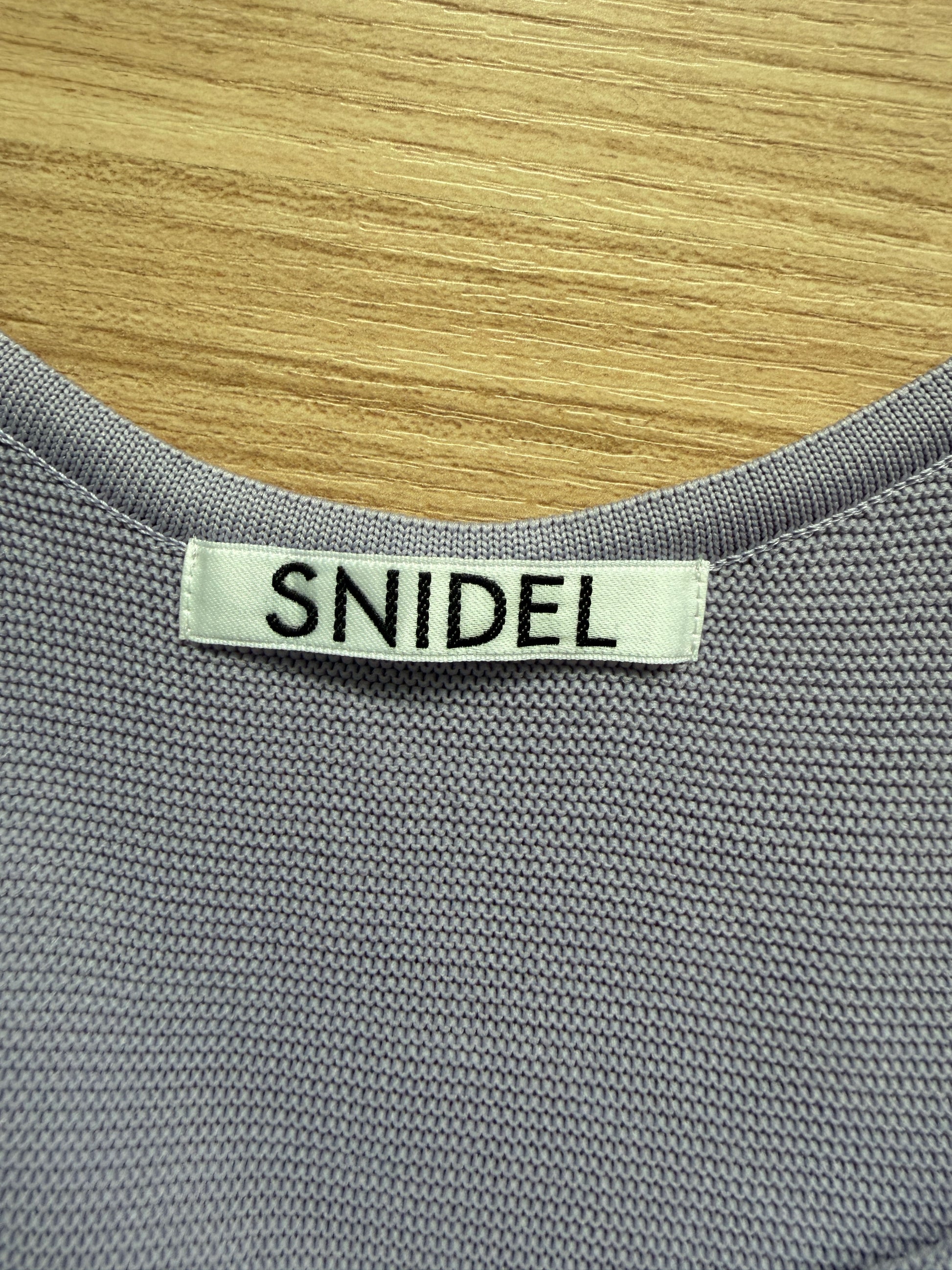 Close-up of a gray ribbed knit garment with a white fabric neck label reading 'SNIDEL' in black text, placed against a light wood background.