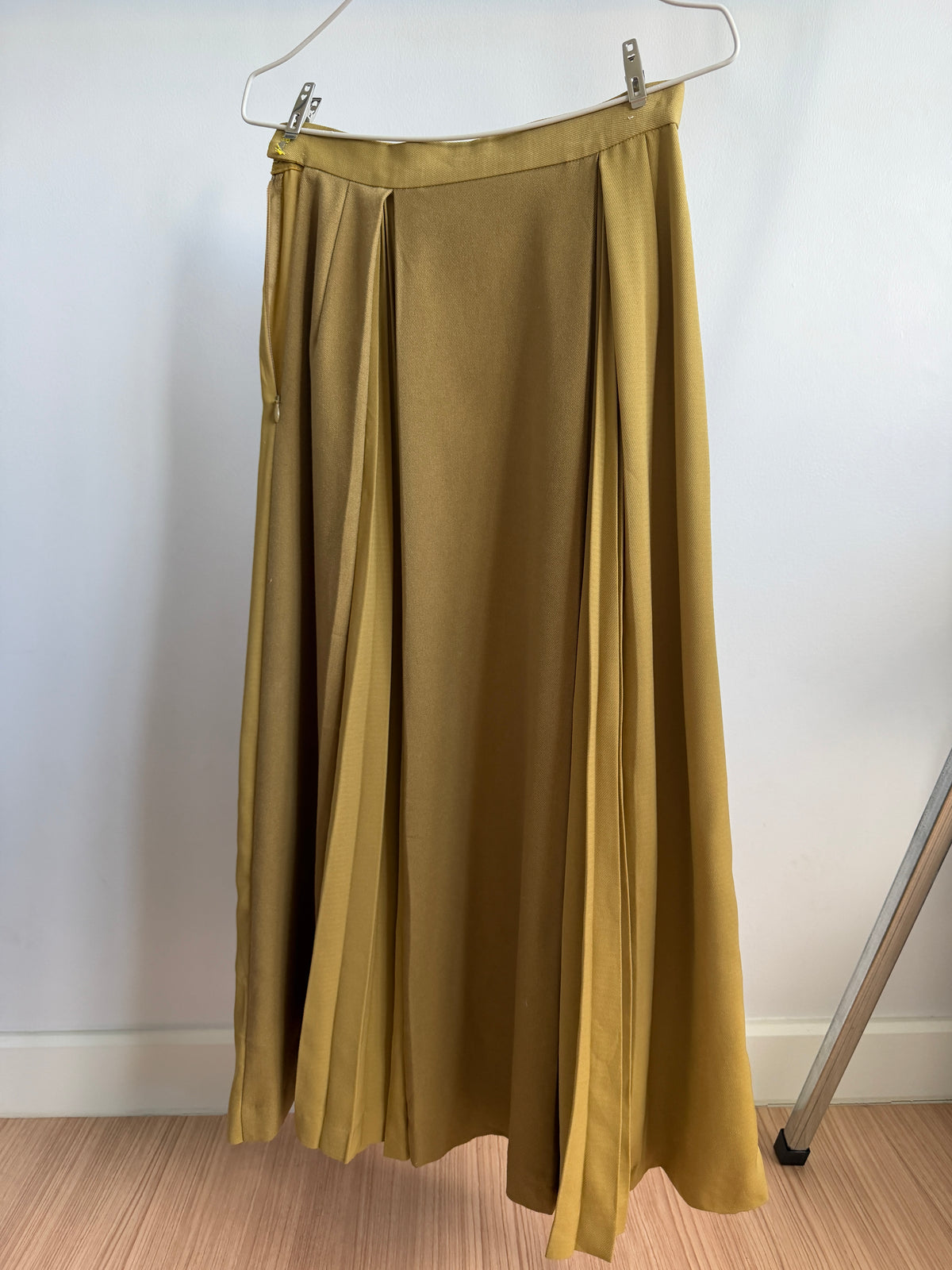 A pleated midi skirt in a warm mustard yellow or ochre color, hanging from a white plastic hanger against a plain white wall.