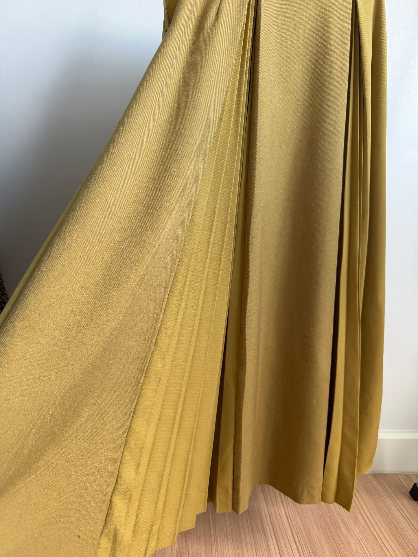A close-up of a pleated mustard yellow fabric, likely a skirt or drape, hanging against a plain white wall and light wood floor.