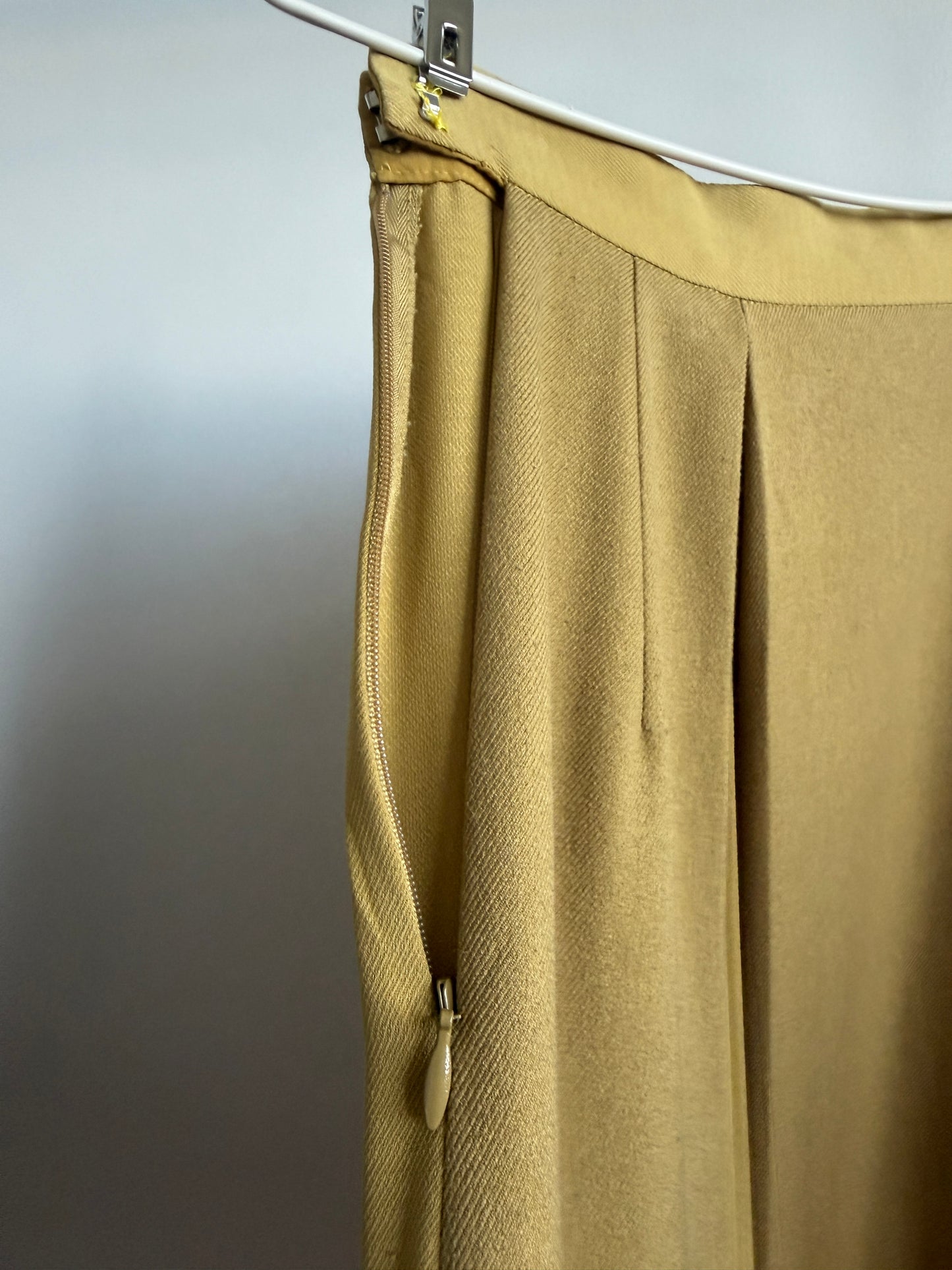 A close-up of a mustard yellow pleated skirt or drape hanging from a white hanger against a plain gray wall.