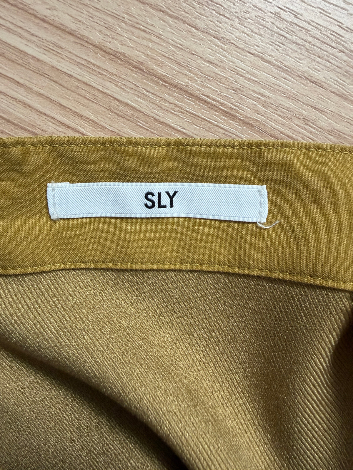 Close-up of a garment's inner neck label reading 'SLY' on a mustard yellow fabric, laid flat on a light wood surface.