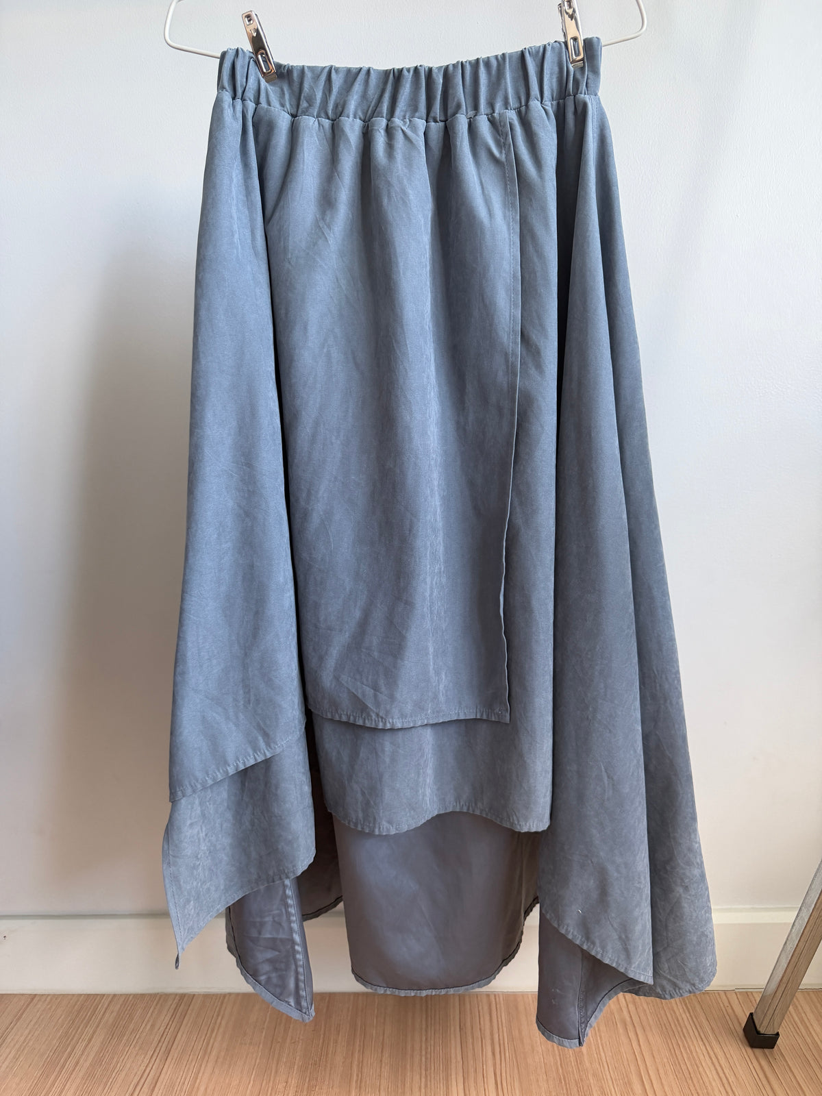 A dusty blue high-low skirt with an elastic waistband, hanging from a metal hanger against a plain white wall on a light wood floor.