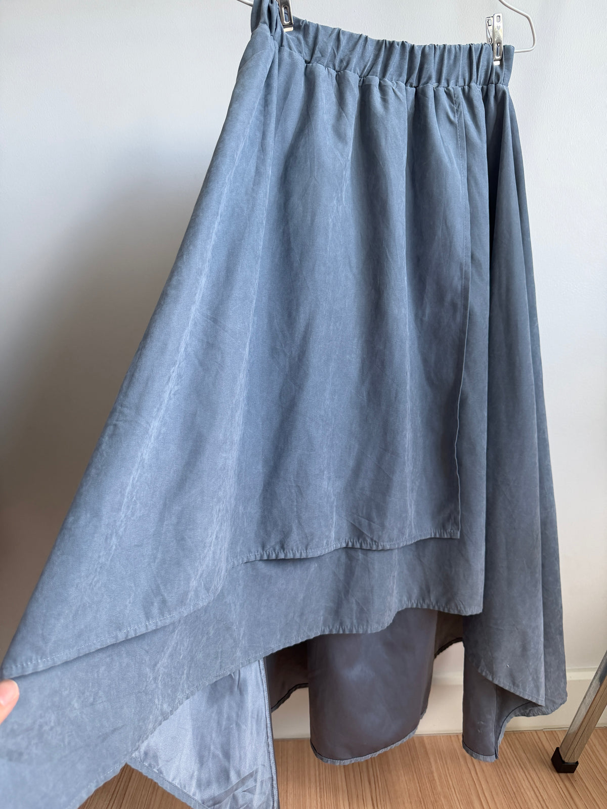 A dusty blue high-low skirt with an elastic waistband, hanging on a metal hanger against a plain wall.