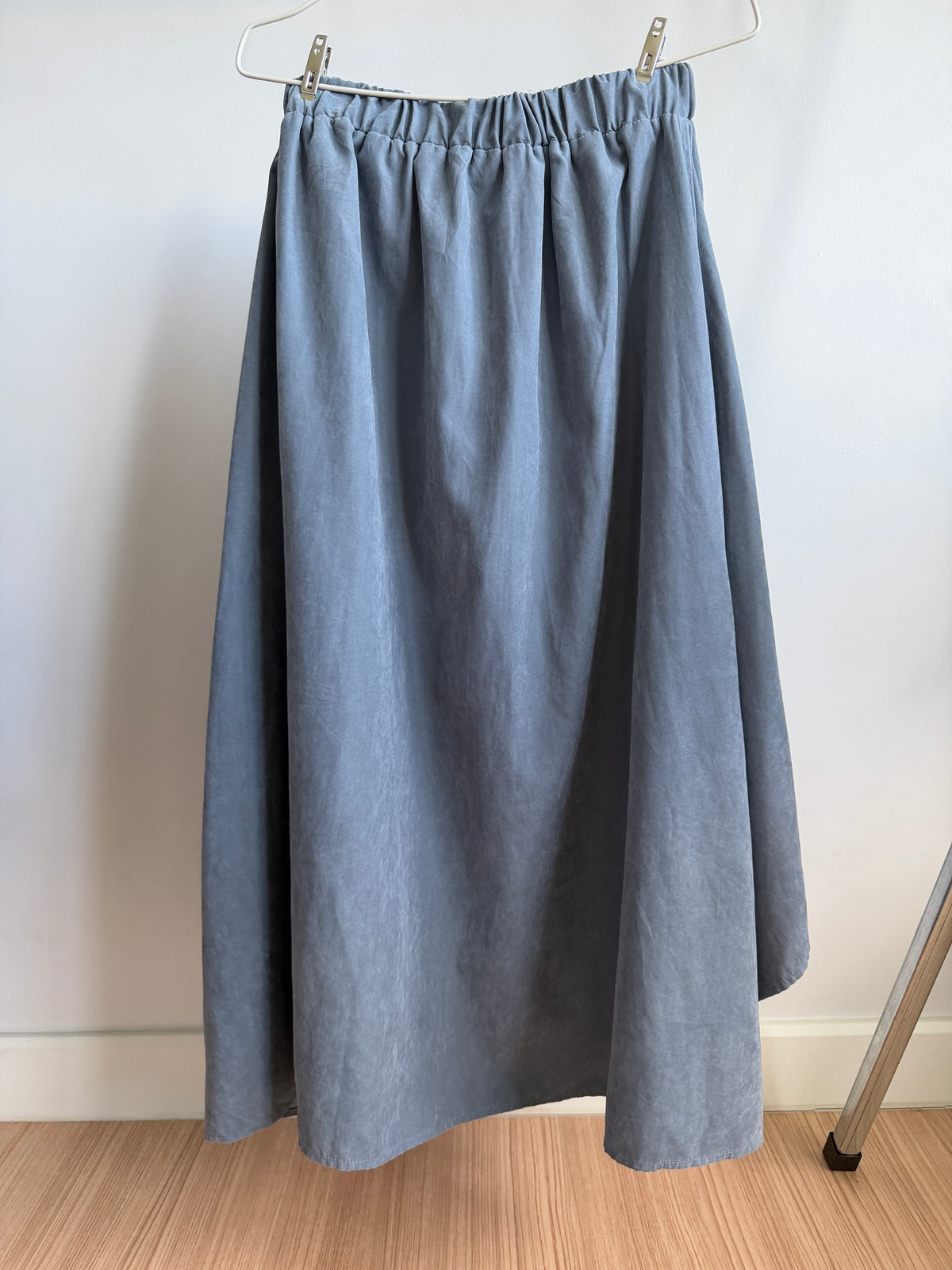 A dusty blue linen midi skirt with an elastic waistband, hanging from a white hanger on a wooden floor against a white wall.